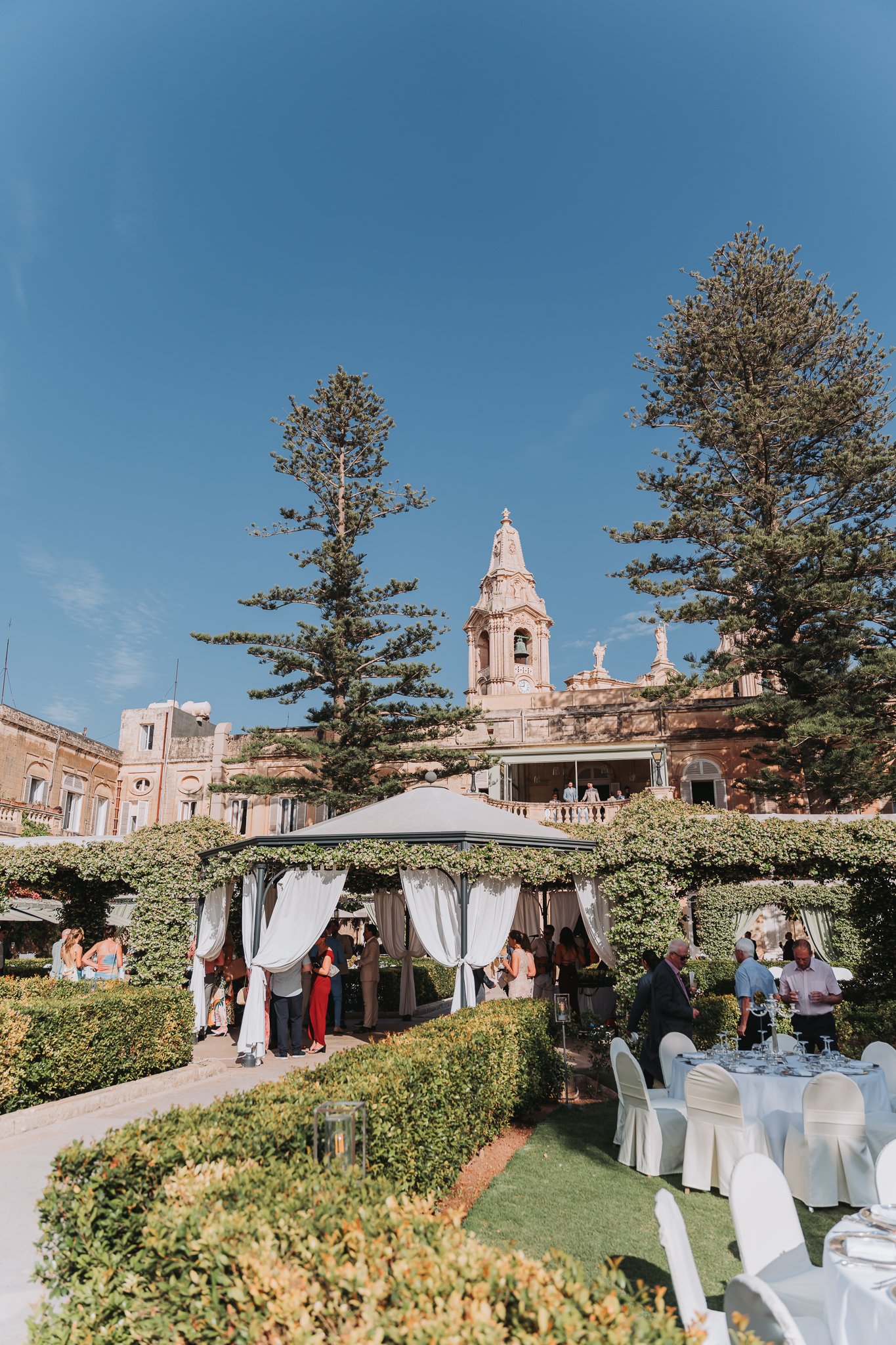 Outdoor wedding reception in a garden with white chairs and tables, white drapes on a gazebo, lush green bushes, tall trees, a historic church tower, and guests mingling under sunny skies.