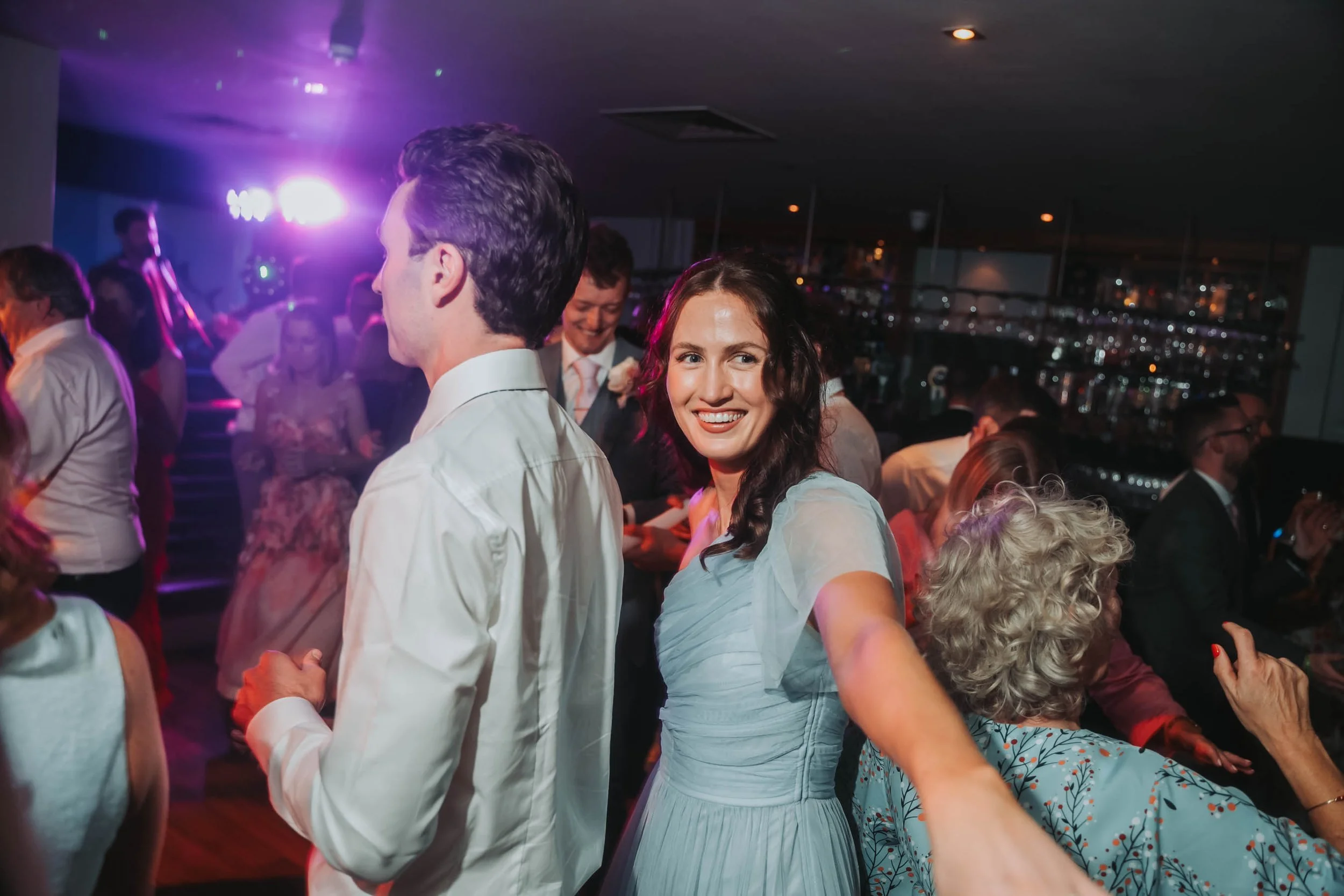 People dancing together at a party or dance event in a dimly lit room with colorful lights.