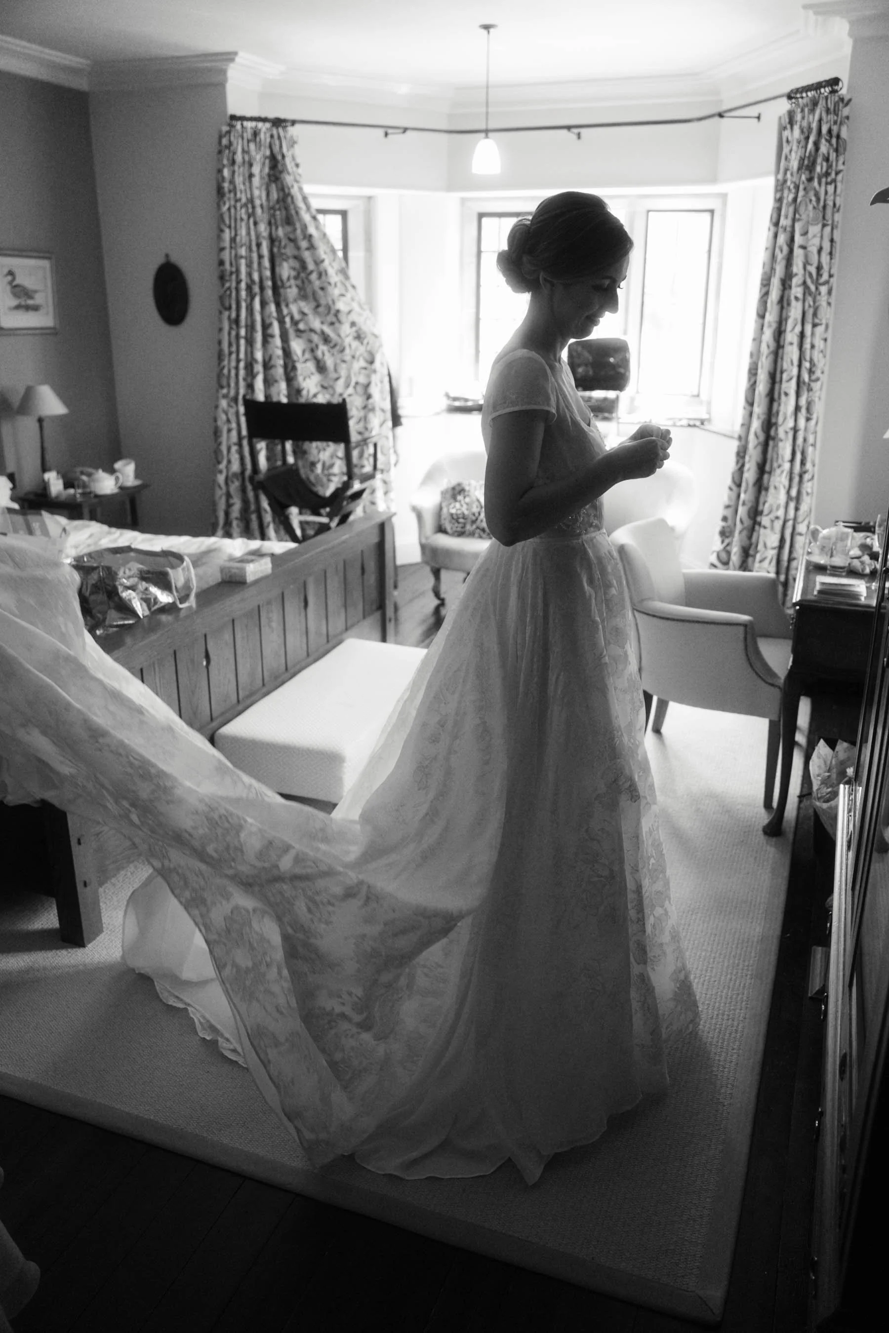 A woman in a wedding dress standing inside a well-lit room, adjusting her dress with a large window and curtains behind her.