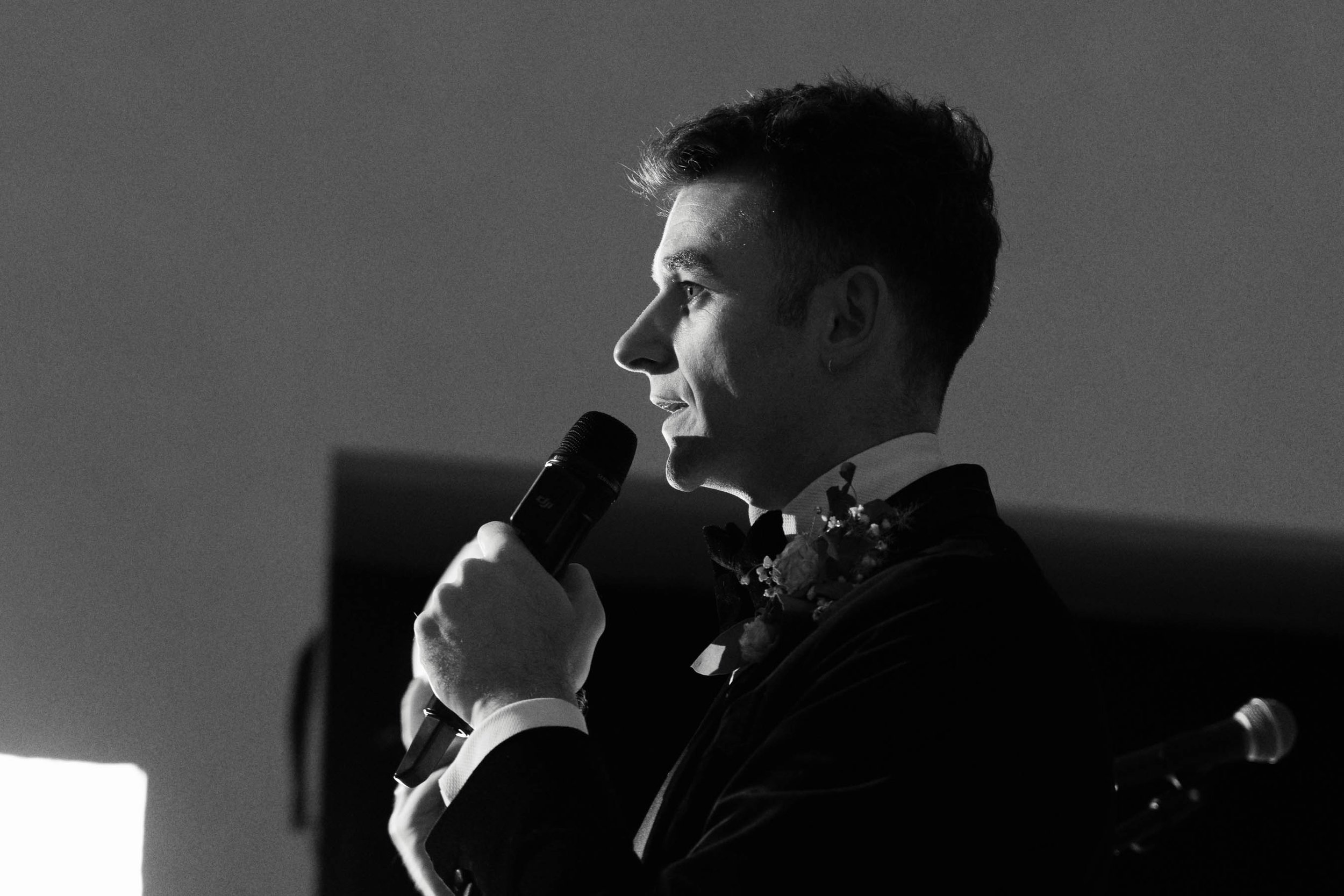 A black and white photo of a young man in a tuxedo holding a microphone, possibly giving a speech or singing, with a bouquet of flowers on his lapel.