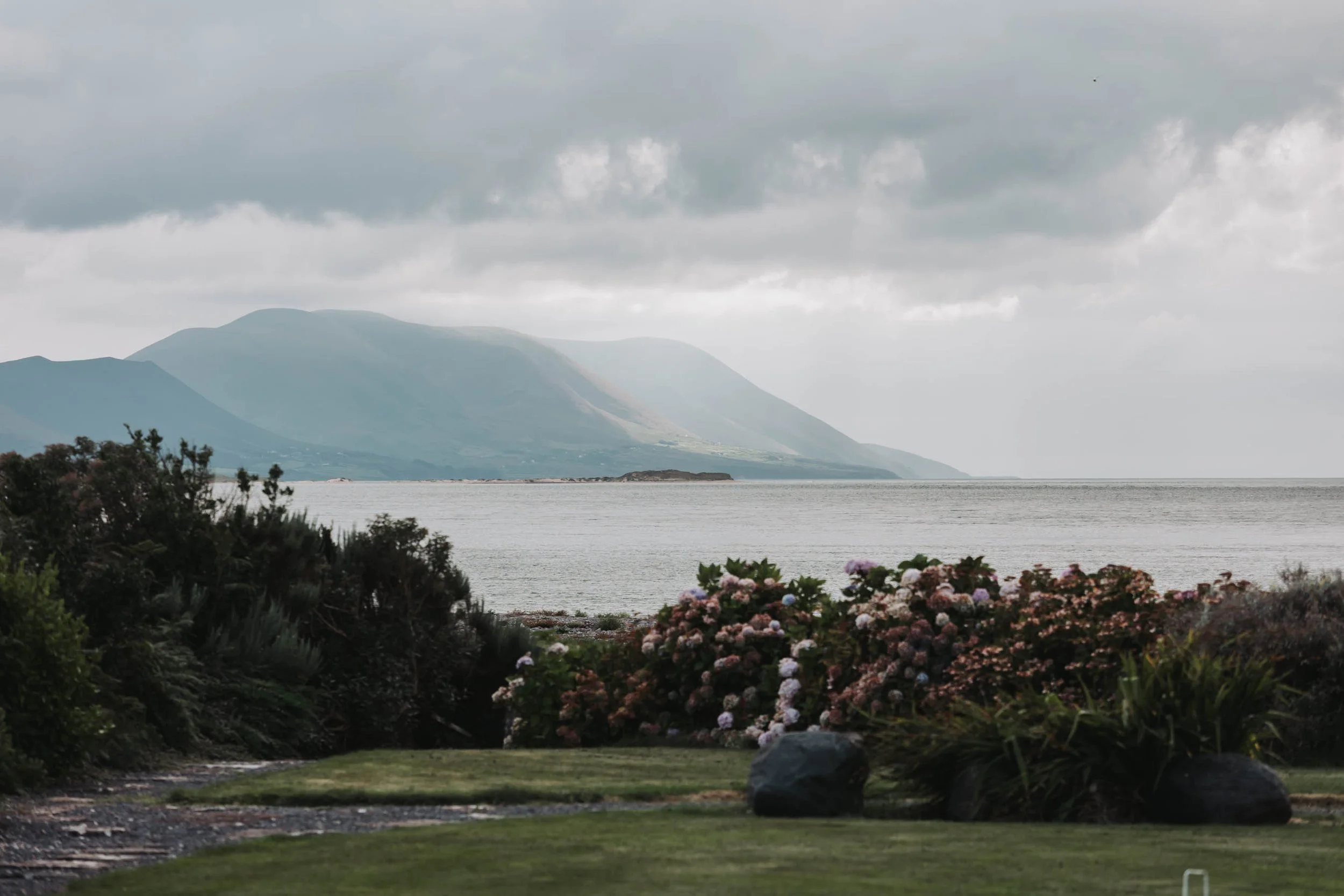 A scenic view of a lake with mountains in the background, cloudy sky, and a garden with blooming flowers in the foreground.