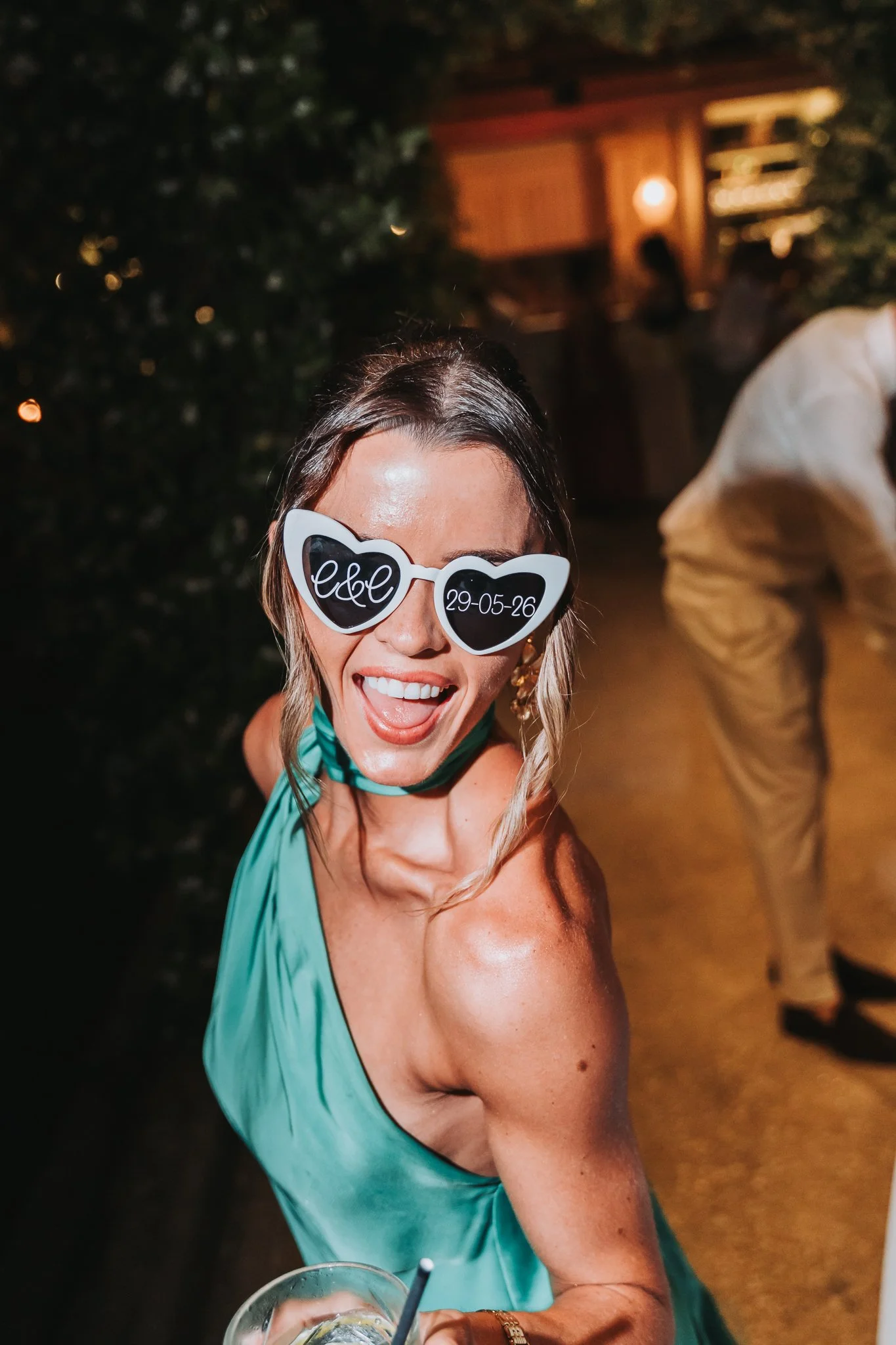 A woman smiling and wearing large heart-shaped sunglasses with the initials 'ellc' and the date '29-05-26'. She is dressed in a teal dress and holding a drink with ice and lemon. The background is dark with some wood-paneled structure and other peopl