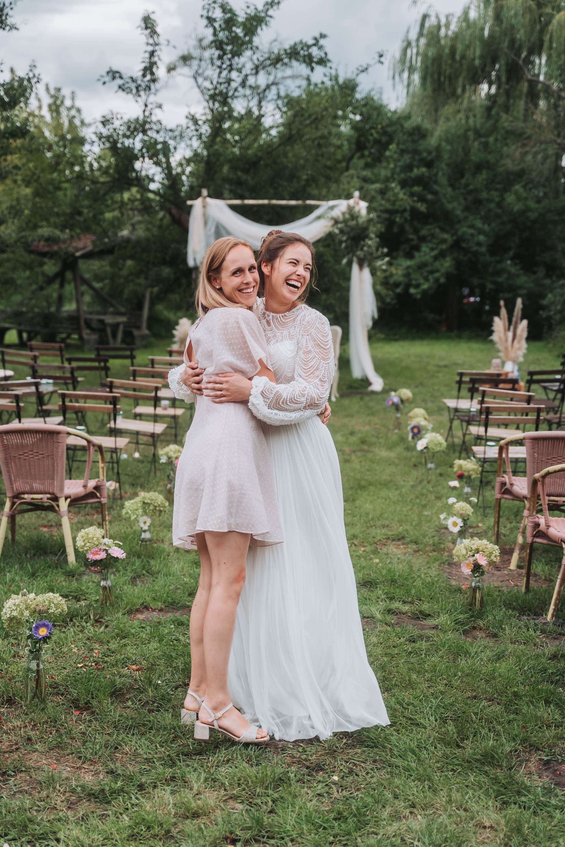 Two women in wedding dresses embracing each other and smiling at a wedding ceremony outdoors. Wooden chairs and floral arrangements are visible, with a decorated arch in the background surrounded by trees.