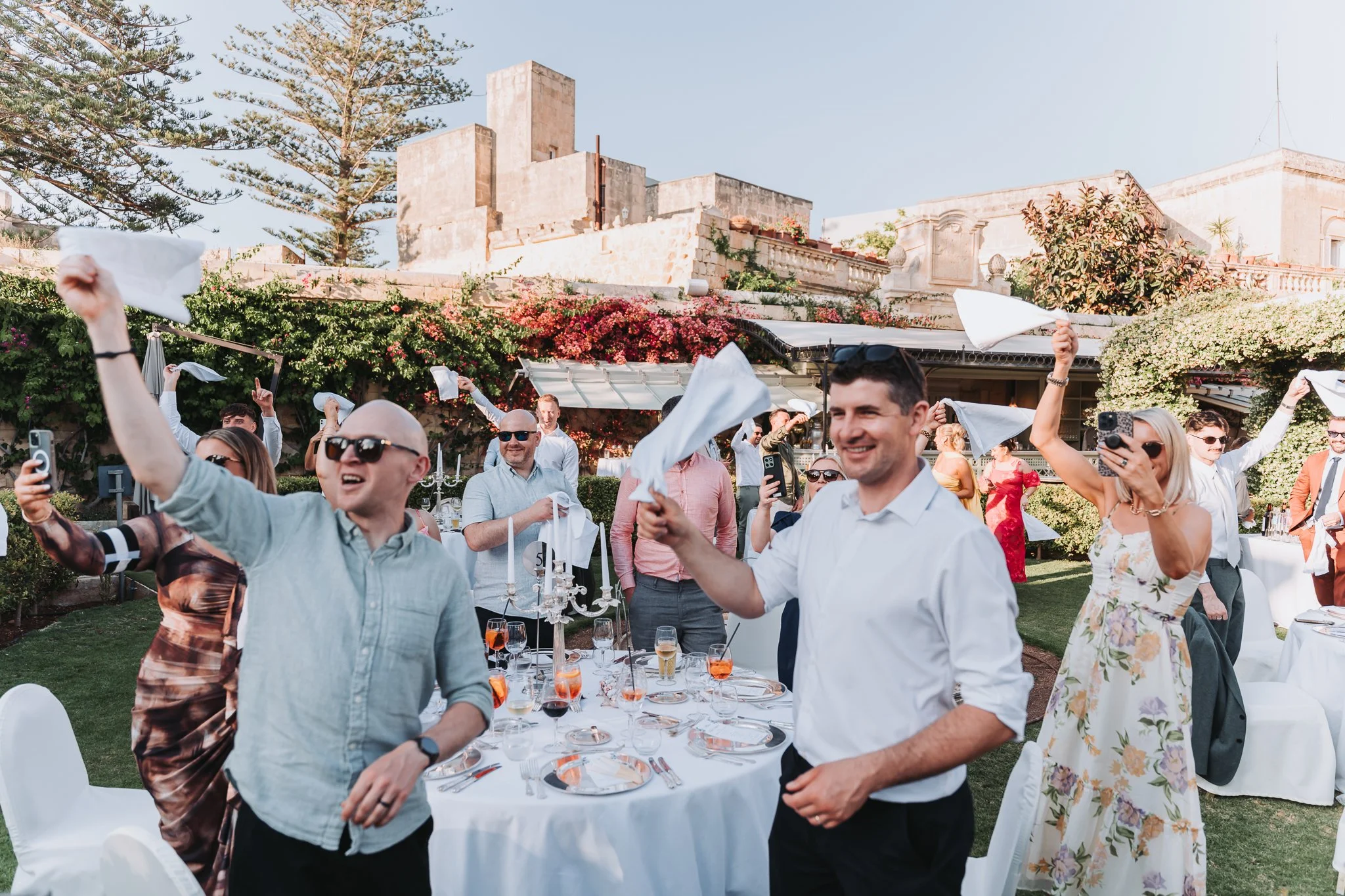 People celebrating outdoors at a party, waving napkins in the air, with a table set with glasses and plates in front of them, and flowering bushes and buildings in the background.