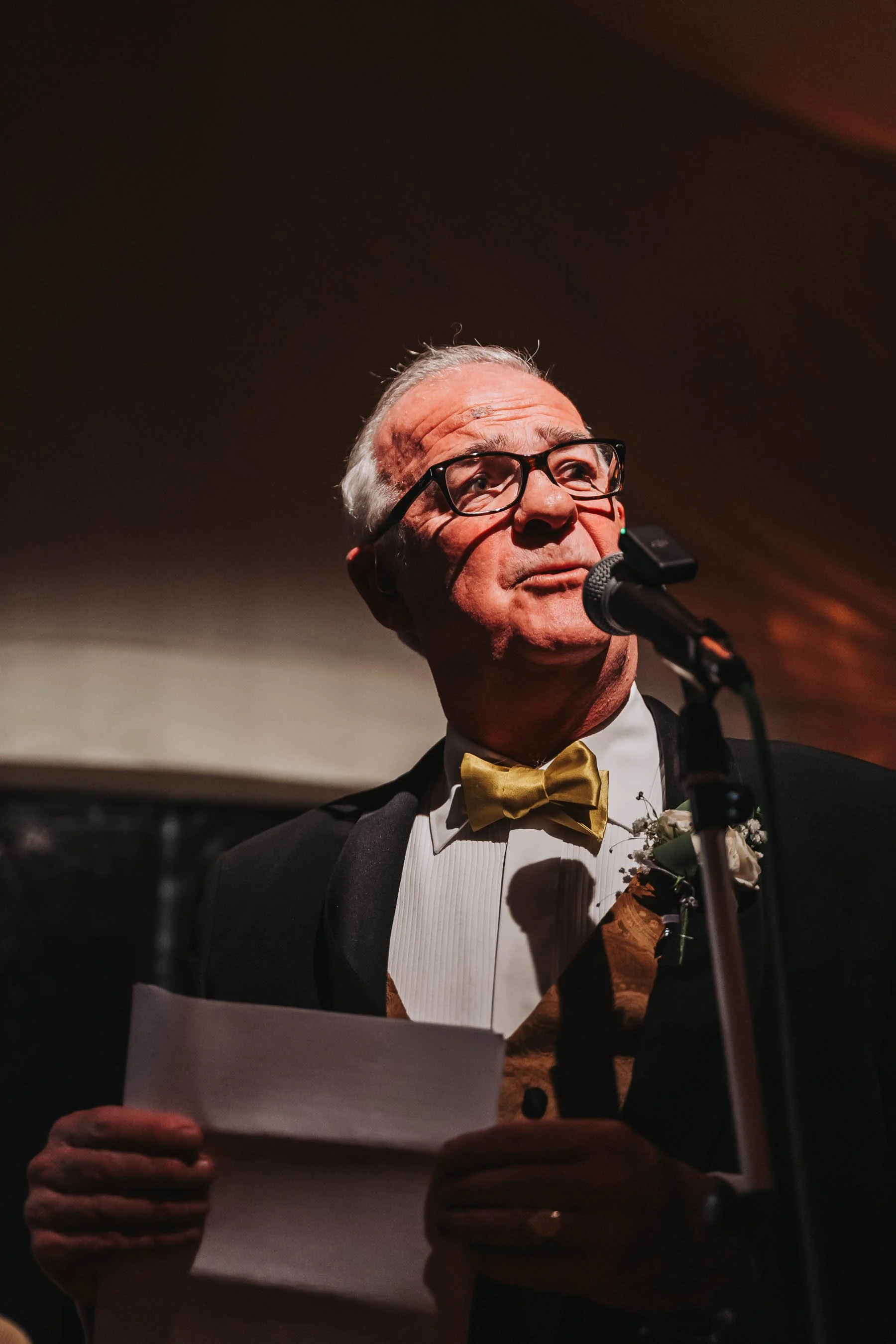 An older man wearing glasses and a tuxedo with a gold bow tie and floral boutonniere, speaking into a microphone while holding a piece of paper at a formal event.