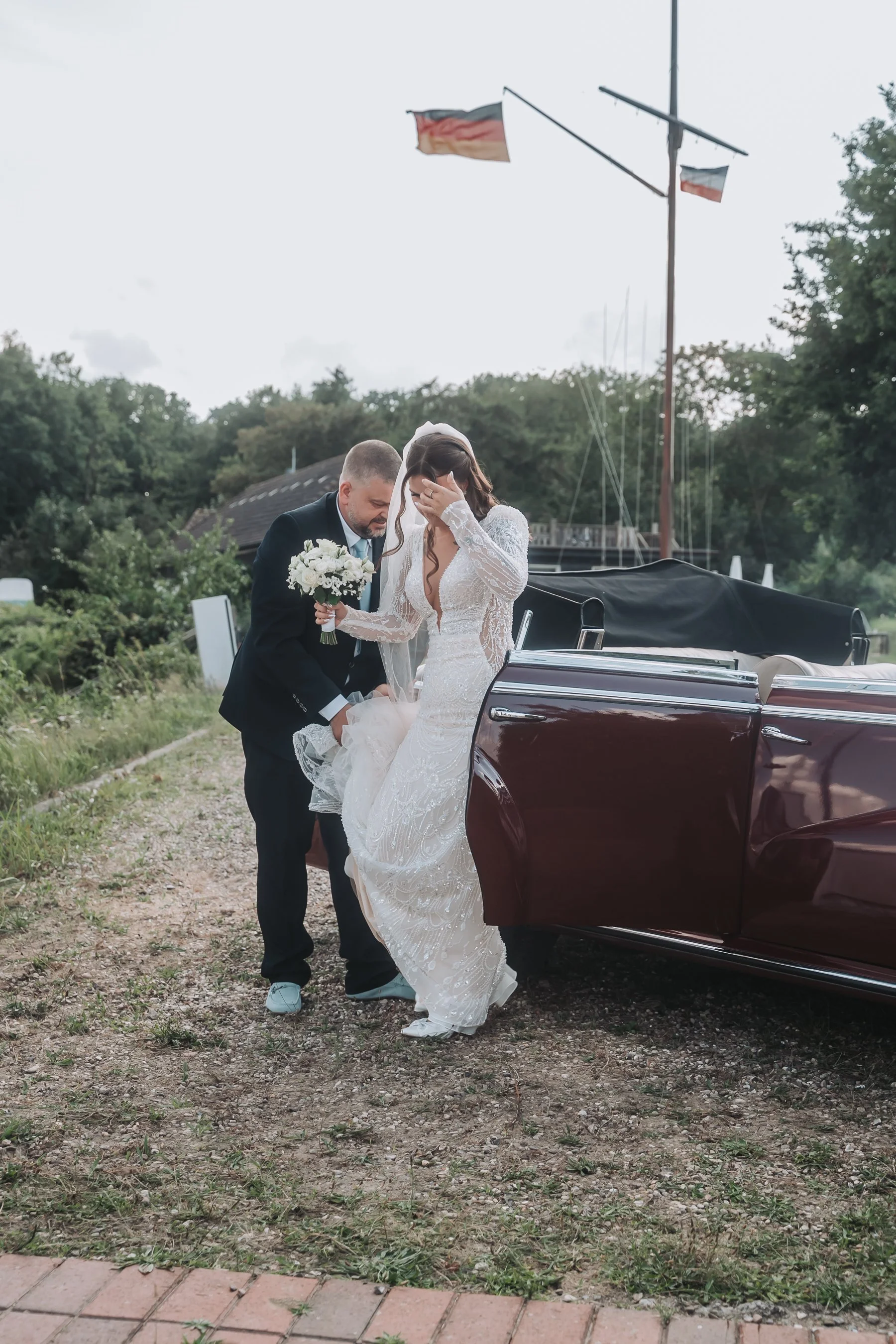 A bride and groom are standing next to a maroon convertible car outside, with the groom helping the bride with her gown. The bride is holding a bouquet of white flowers, and both are dressed in wedding attire. The background includes trees, a flagpol