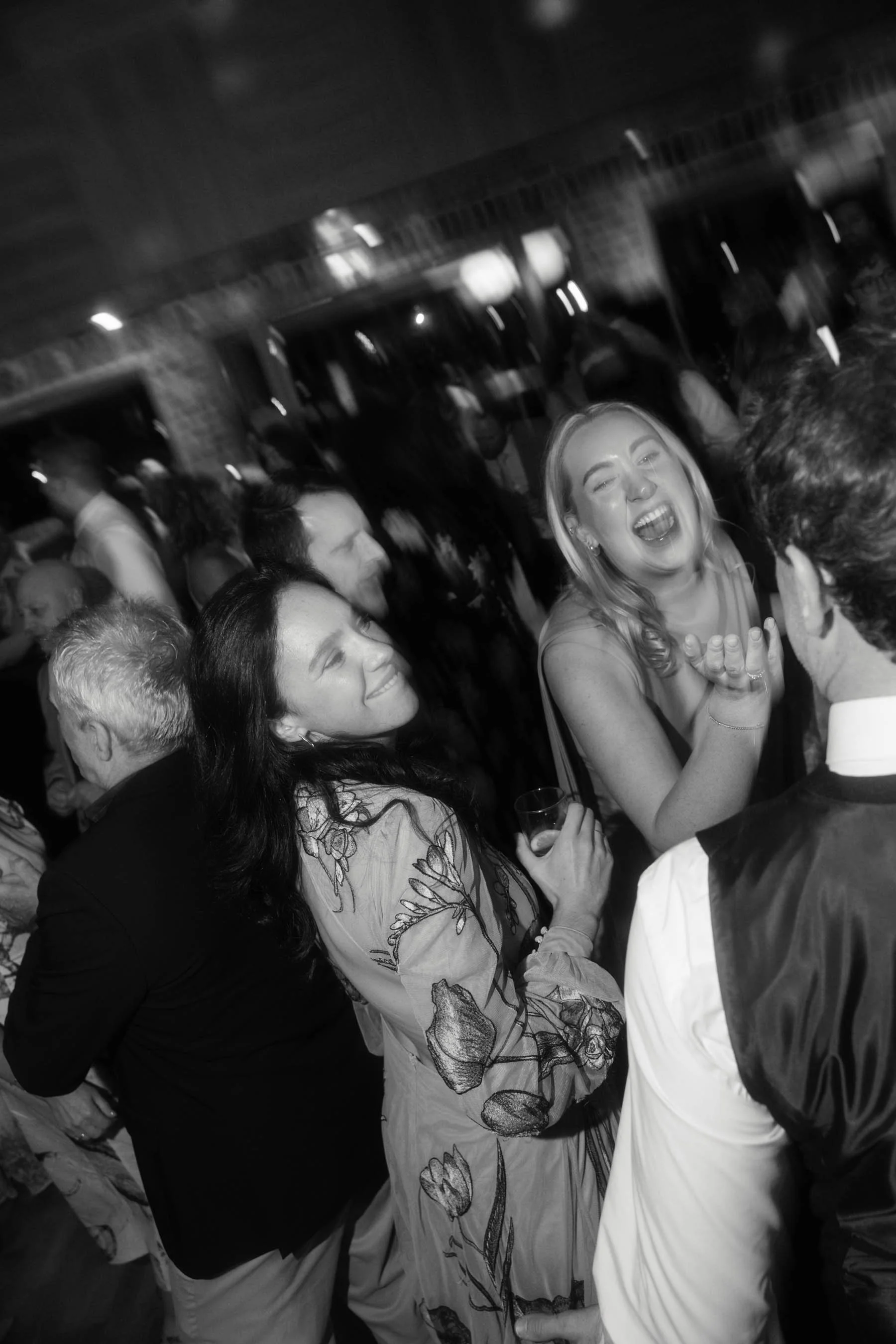 A black and white photo of a lively party scene with multiple people socializing indoors. The focus is on a smiling woman with long blonde hair holding a glass, surrounded by others in conversation and laughter.