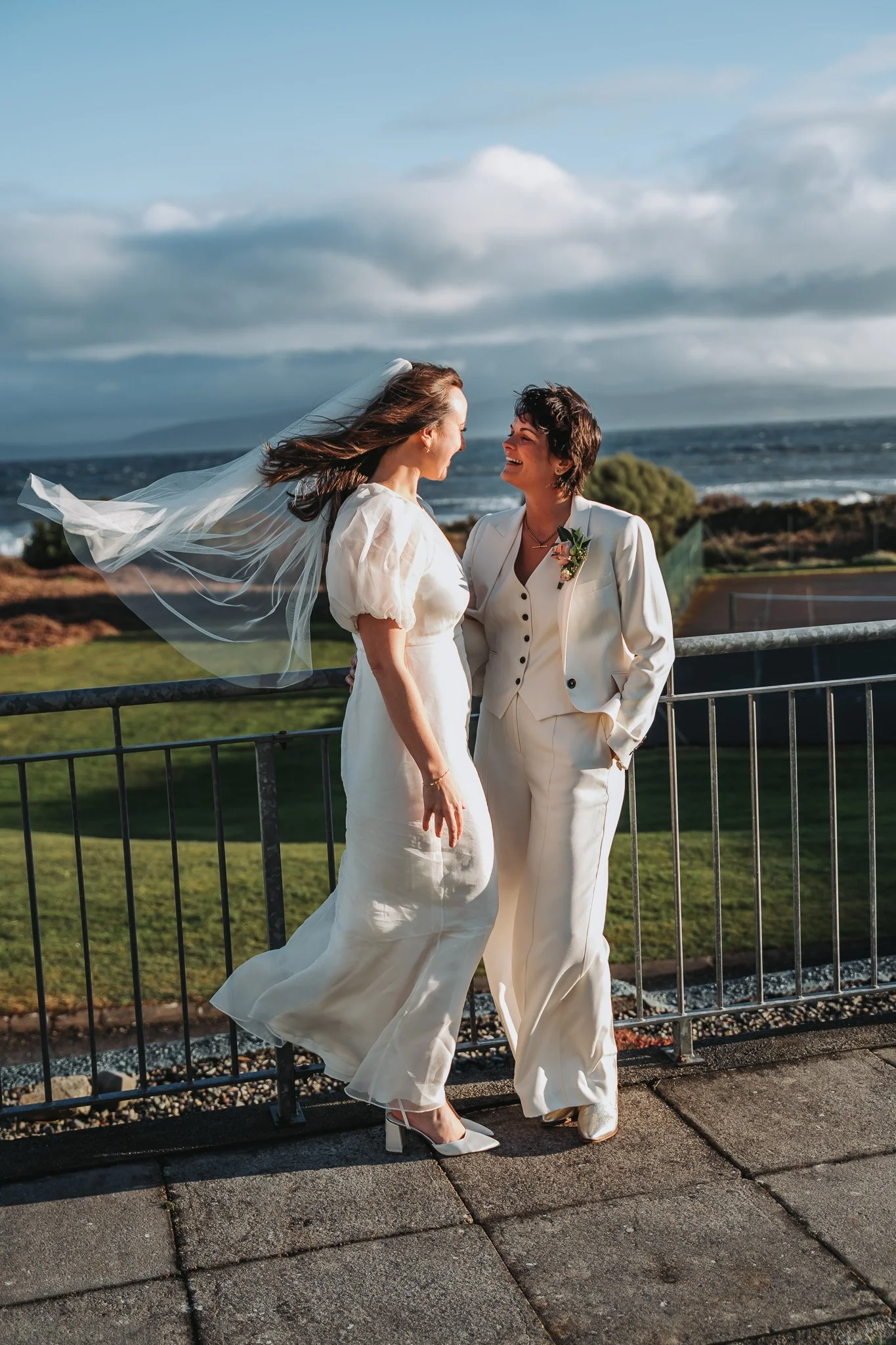 Two women, one in a wedding dress with a veil and the other in a cream suit, sharing a joyful moment outdoors near the ocean with a cloudy sky.