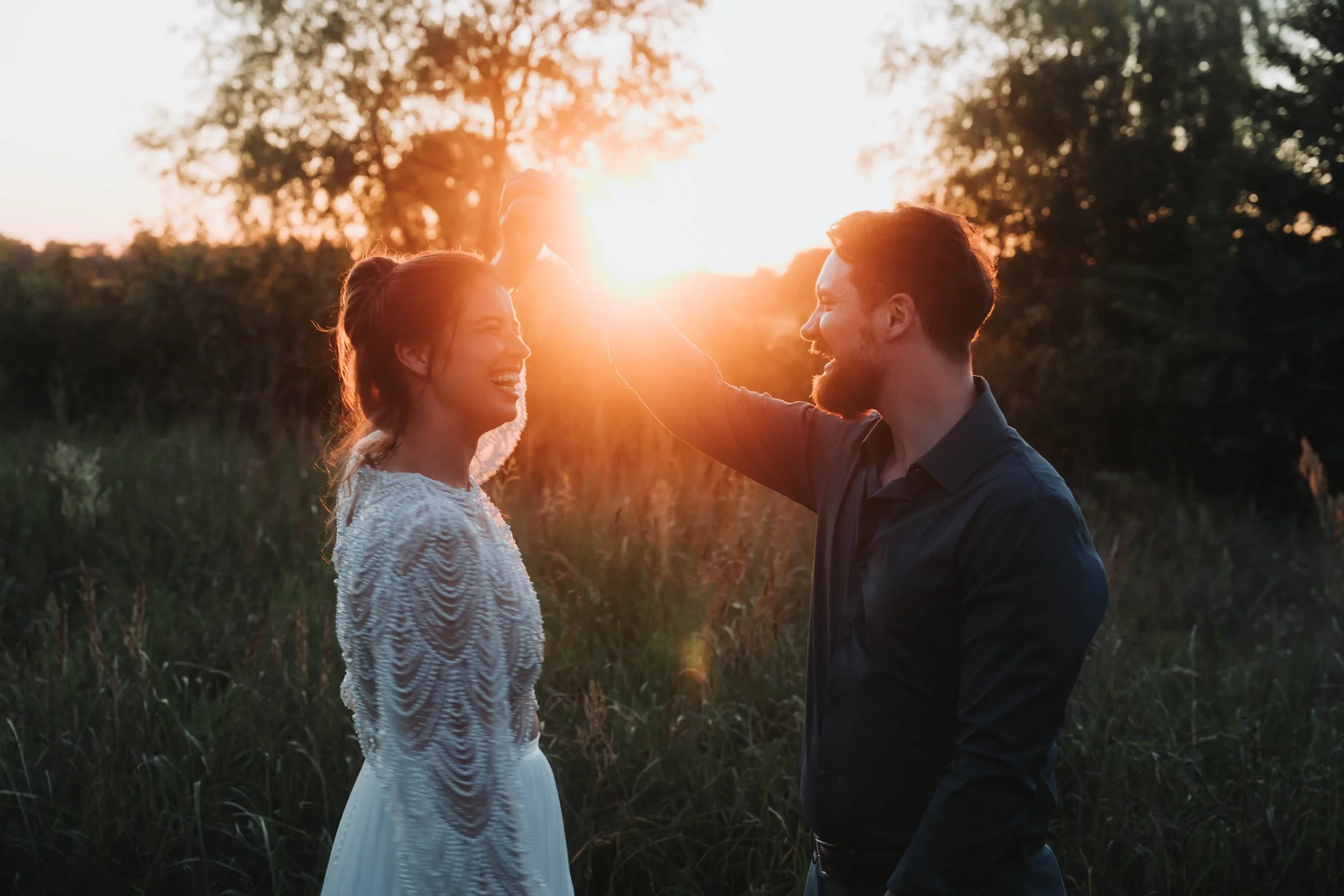A man and a woman are outdoors at sunset, smiling and touching foreheads, with the sun setting behind trees in the background.