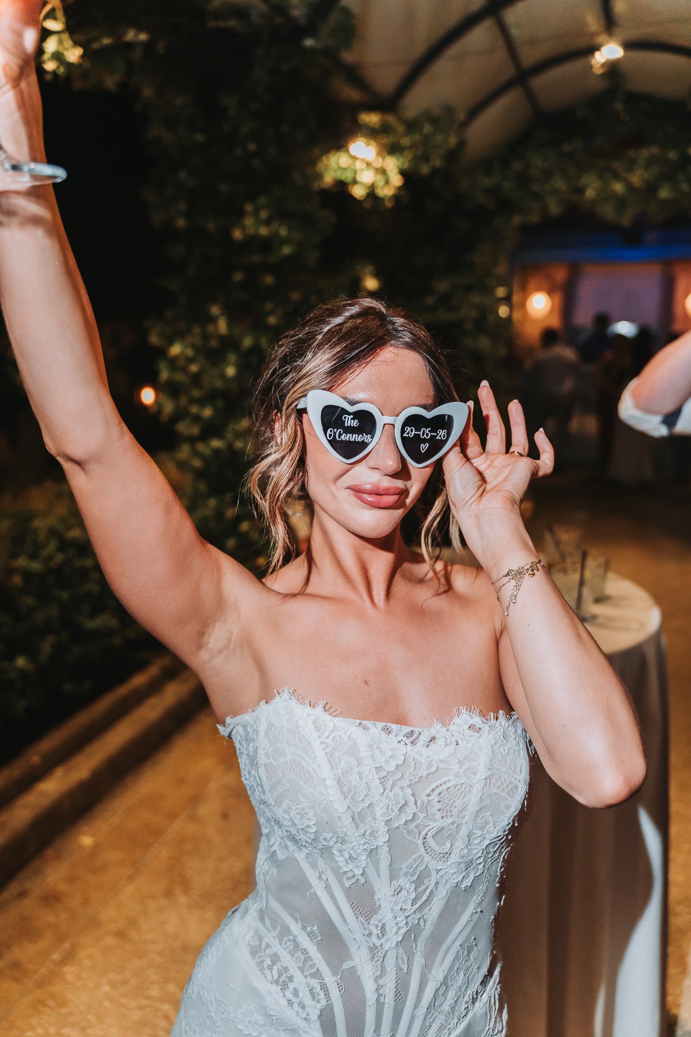 A woman in a white strapless lace wedding dress at an indoor event, wearing white heart-shaped sunglasses with black lenses that read 'The O'Connors 29-05-26' and a small heart graphic, smiling with her eyes closed, with her left hand near her glasse