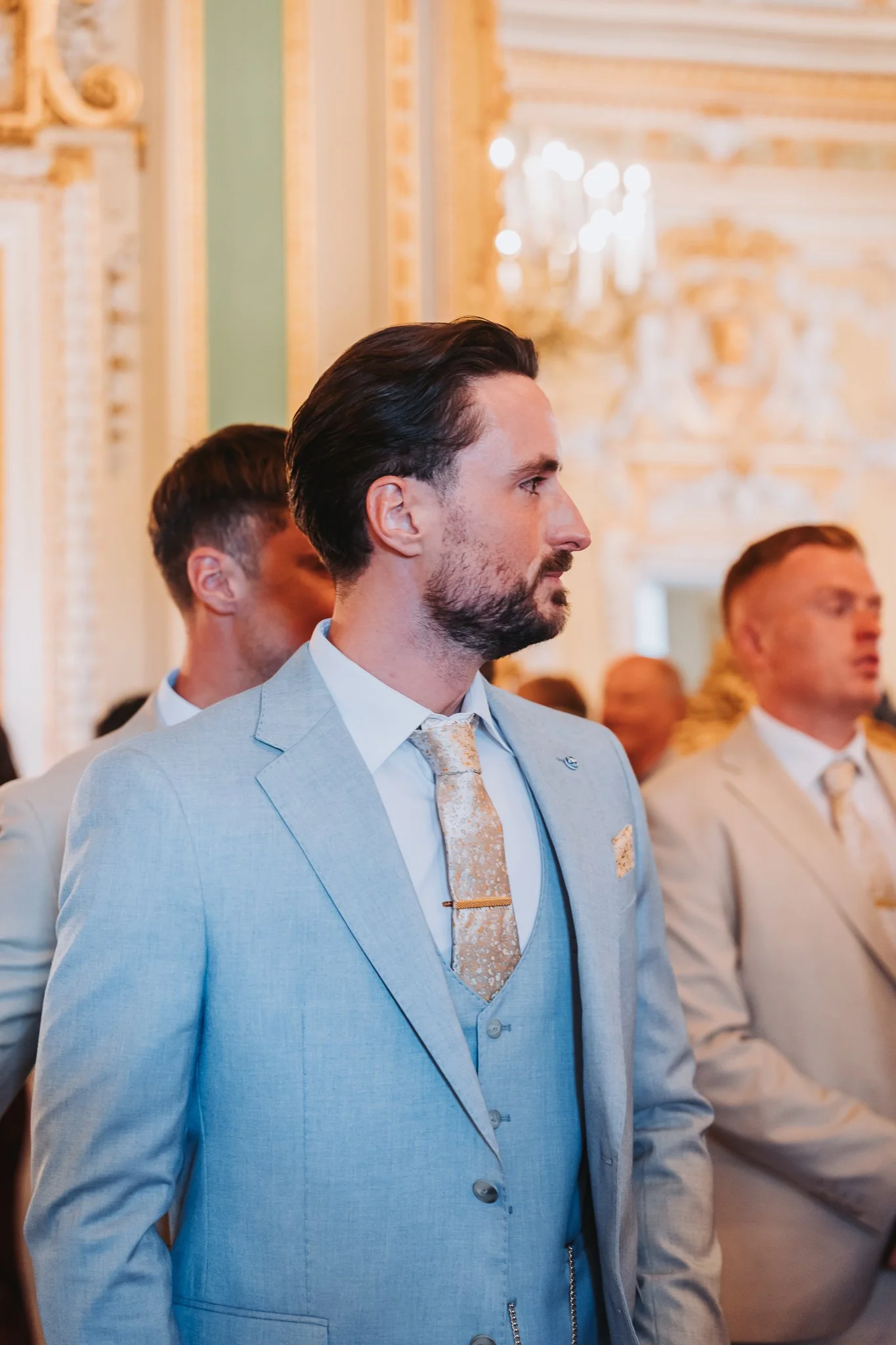 Man with dark hair and a beard wearing a light gray suit with a patterned gold tie, standing in a decorated interior, likely at a formal event.