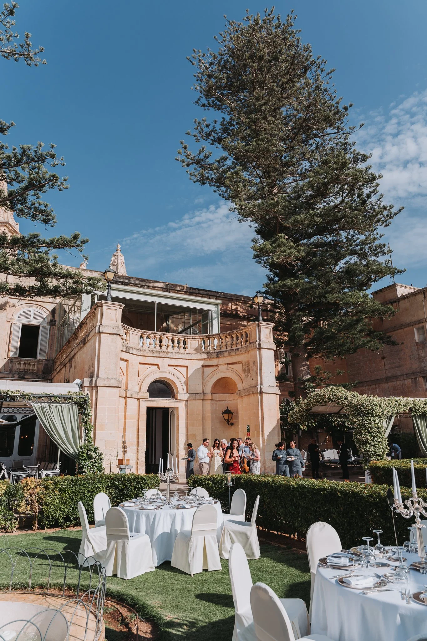 Outdoor wedding reception with round tables covered with white tablecloths and surrounded by white chairs, set in a lush garden with green grass and trees, in front of a historic building with arched windows and ornate architecture. Gathered guests a