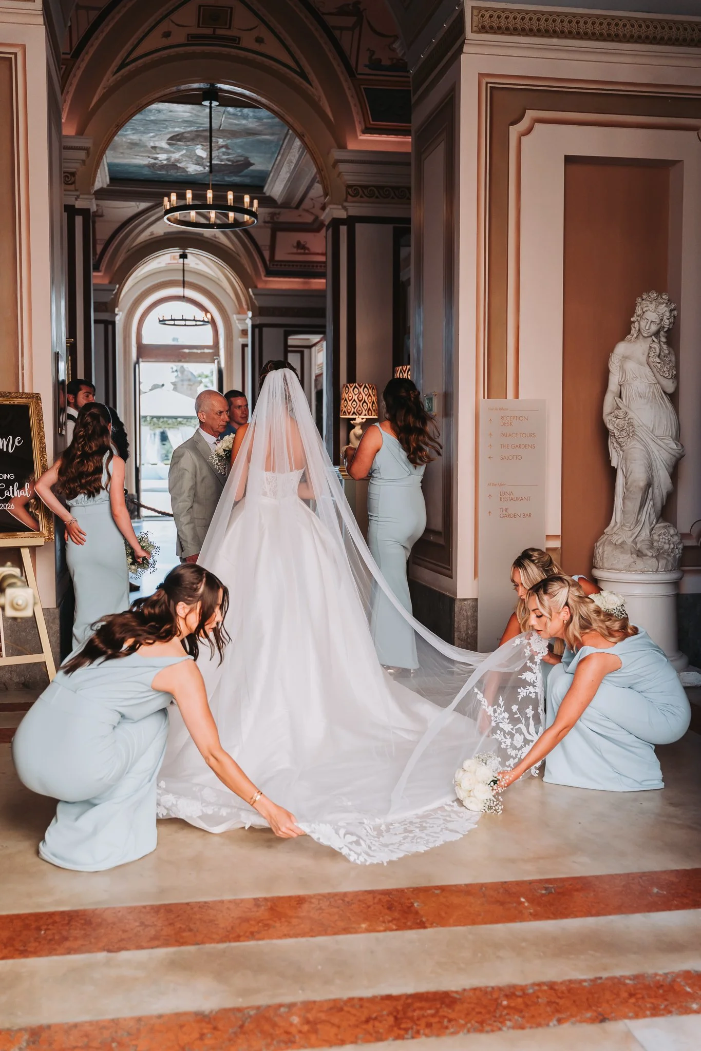 A bride in a wedding gown and veil is surrounded by bridesmaids in light blue dresses, all adjusting the train of the bride's dress inside a decorated hall with murals and statues.