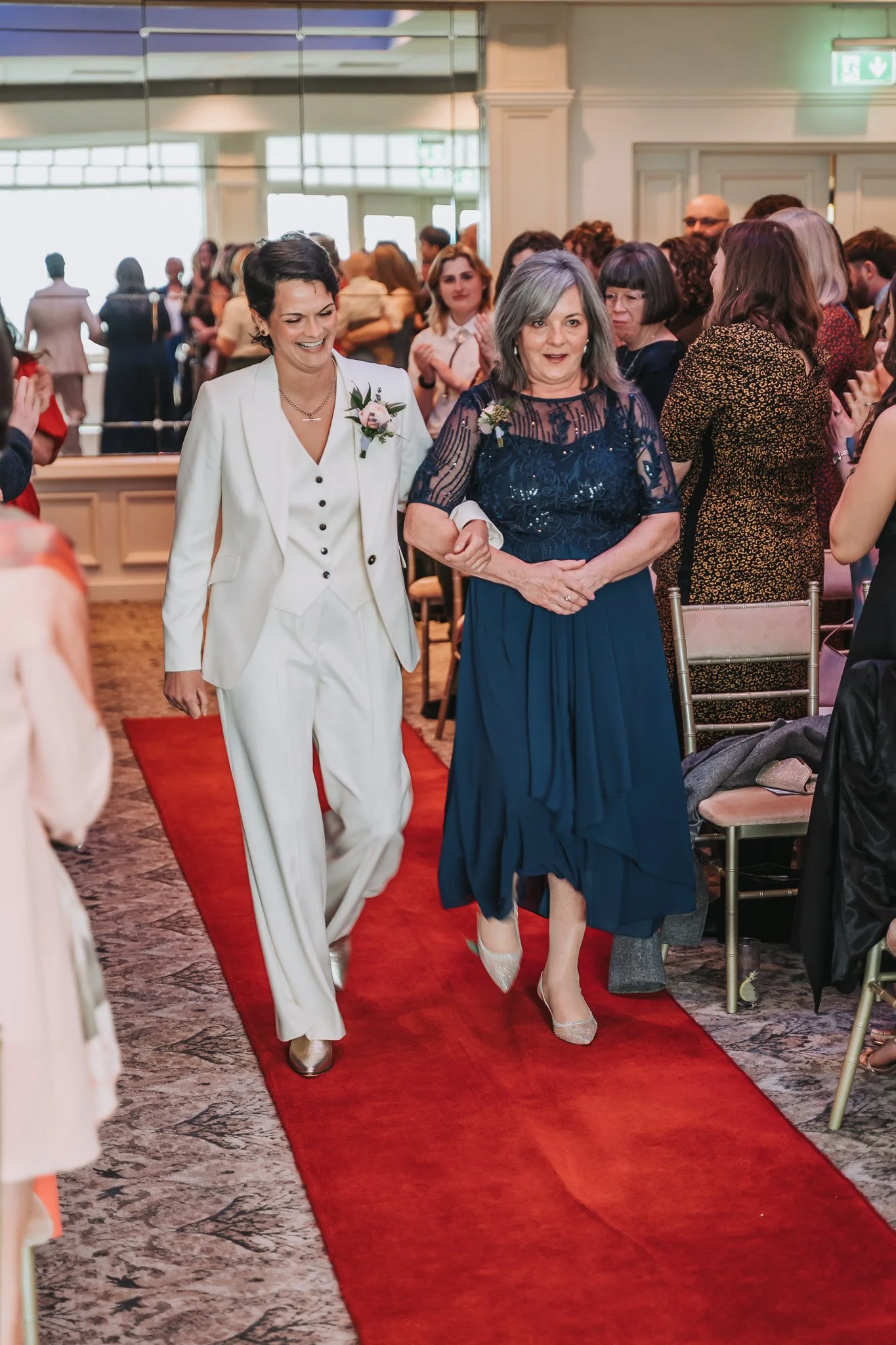 Two women walking down a red carpet at a formal event, surrounded by guests clapping and smiling.