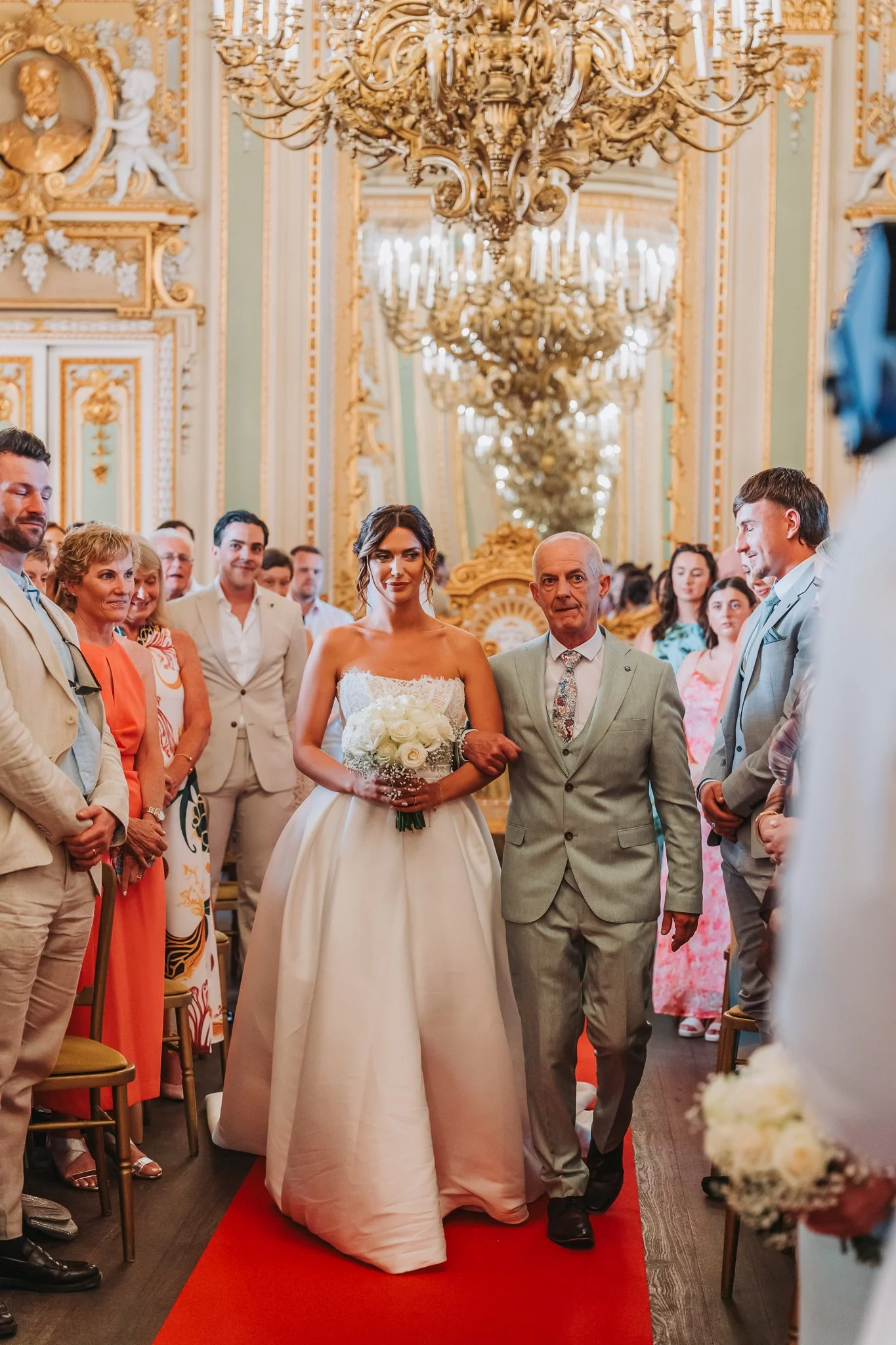 A bride walking down the aisle with her father in a grand, ornate room with chandeliers and gilded decoration, surrounded by wedding guests.