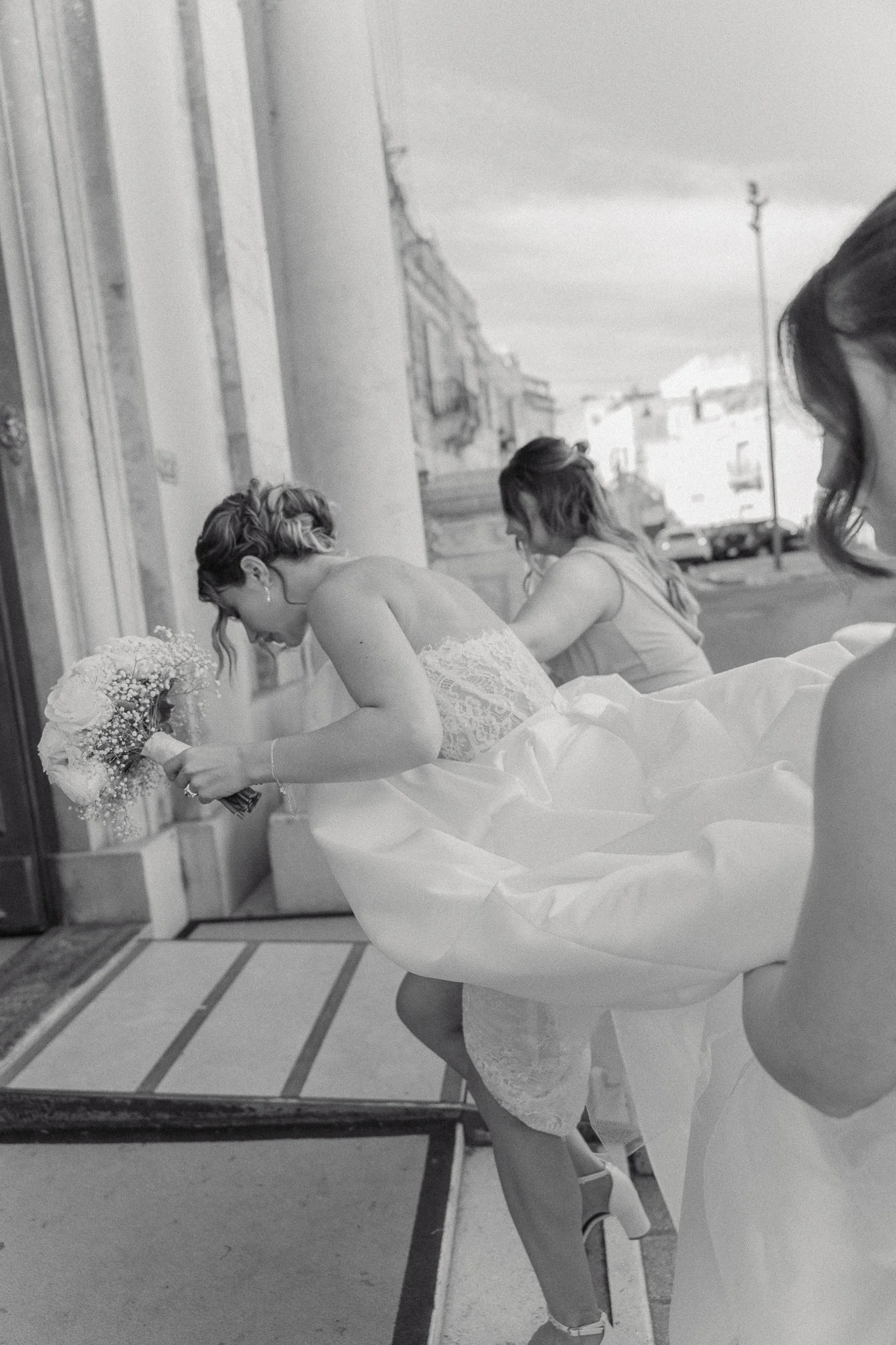 A woman in a wedding gown placing a bouquet of flowers on a ledge, with two other women in dresses nearby, outside a building with a street and parked cars in the background.