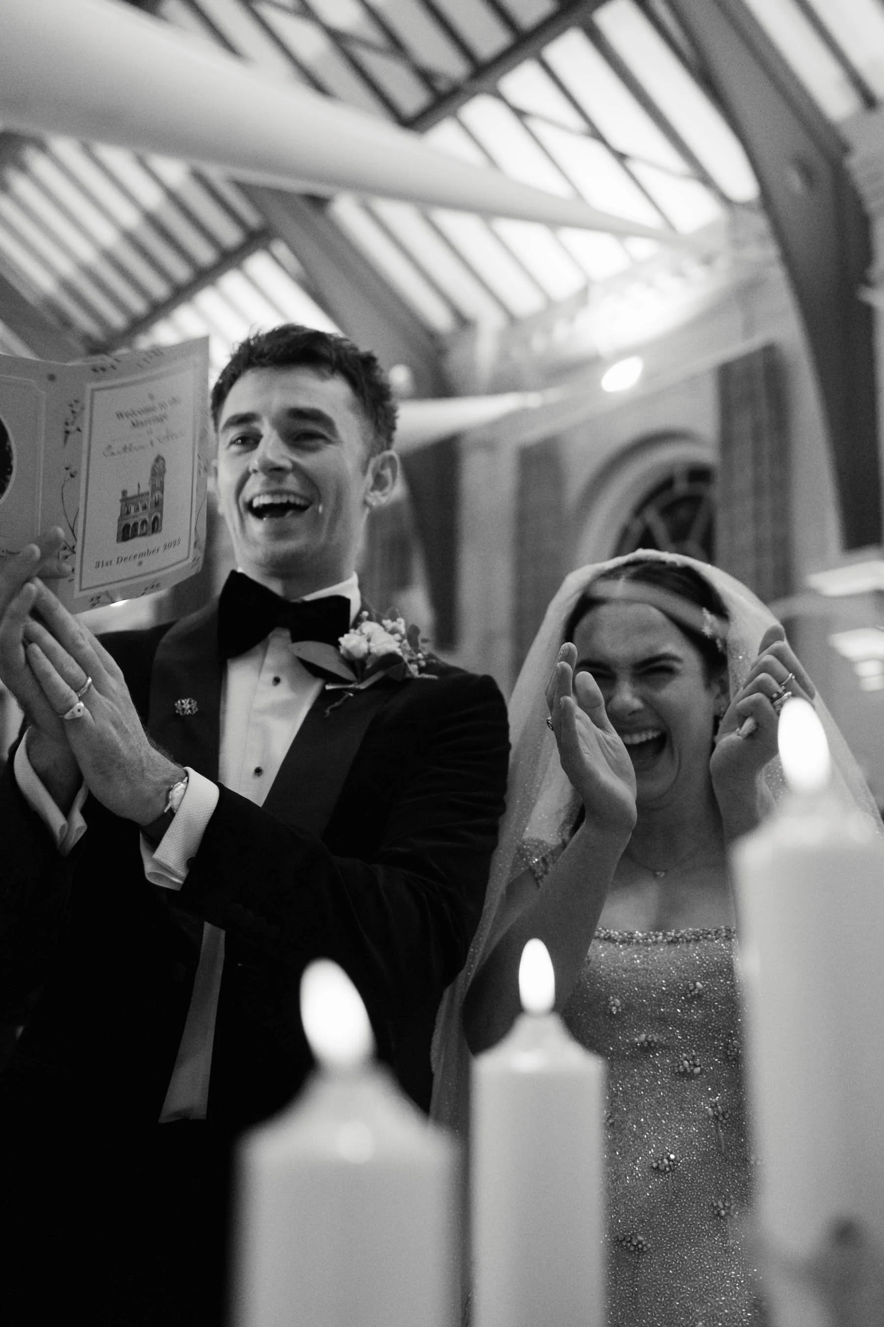 Black and white photo of a smiling young man and woman, dressed formally, likely at a wedding. The man is in a tuxedo, the woman in a sparkly dress with a veil. They appear to be happy and holding a booklet. There are lit candles in the foreground an