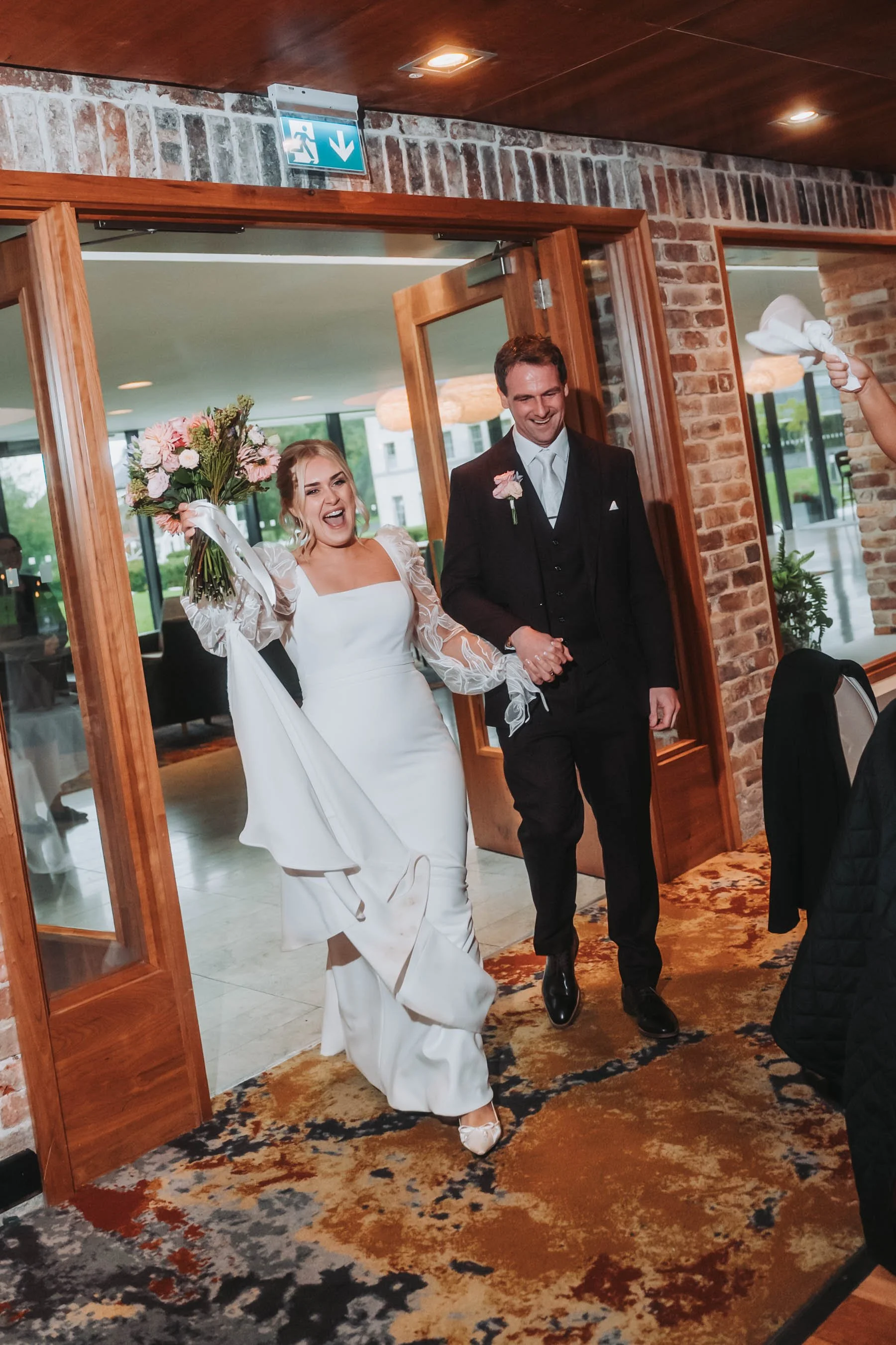 A bride and groom walking through a doorway, holding hands and smiling. The bride is in a white wedding gown and holding a bouquet, while the groom is in a black suit. The setting appears to be a wedding venue with brick walls and glass doors.