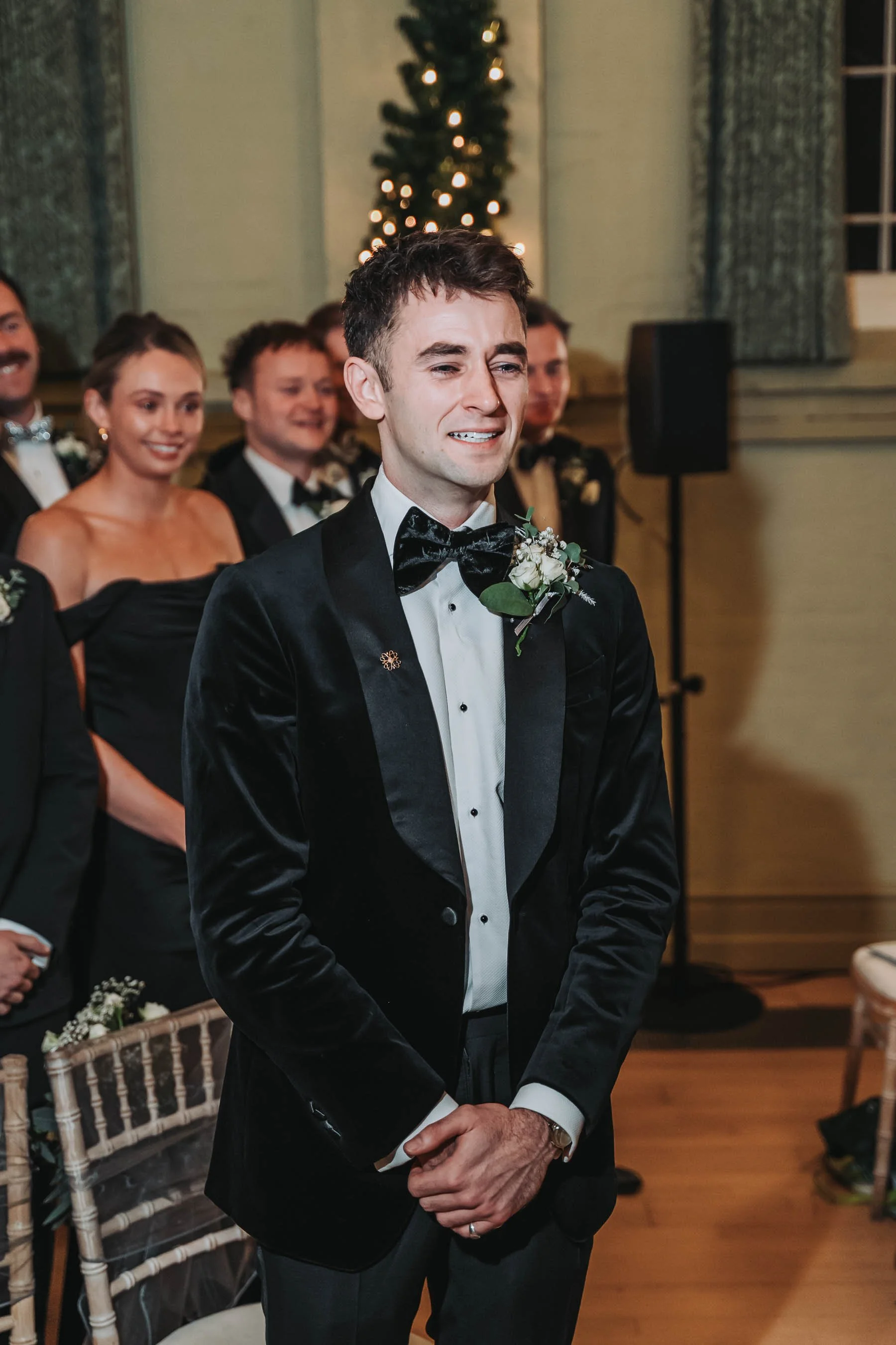 A young man in a black tuxedo with a bowtie and boutonniere, standing with his hands clasped at a formal indoor event, smiling emotionally. In the background, people dressed formally, some smiling, and a decorated Christmas tree can be seen.