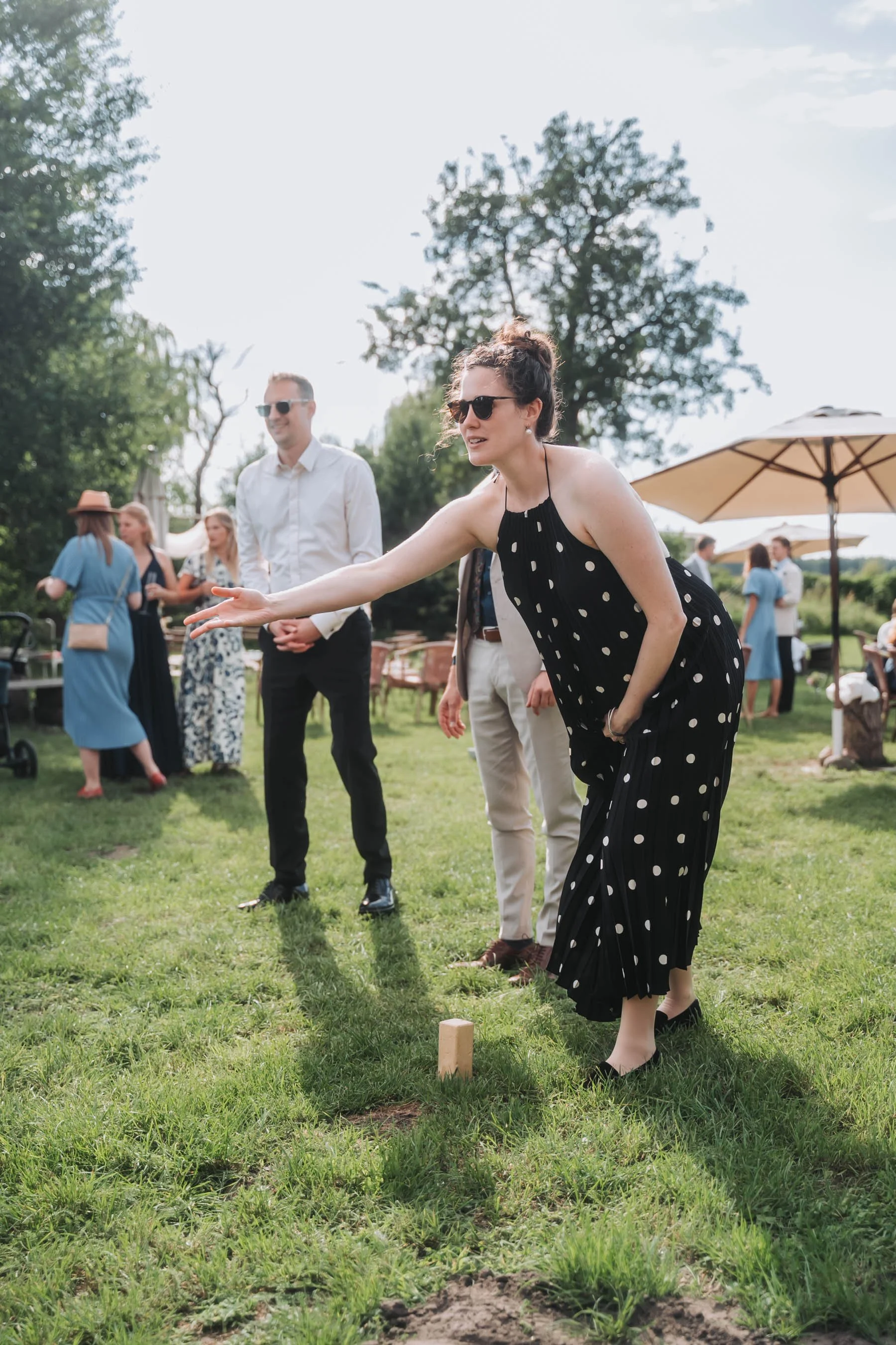 A woman in a black dress with white polka dots throwing a small wooden block in a game of cornhole outdoors at a sunny gathering, with other people and umbrellas in the background.