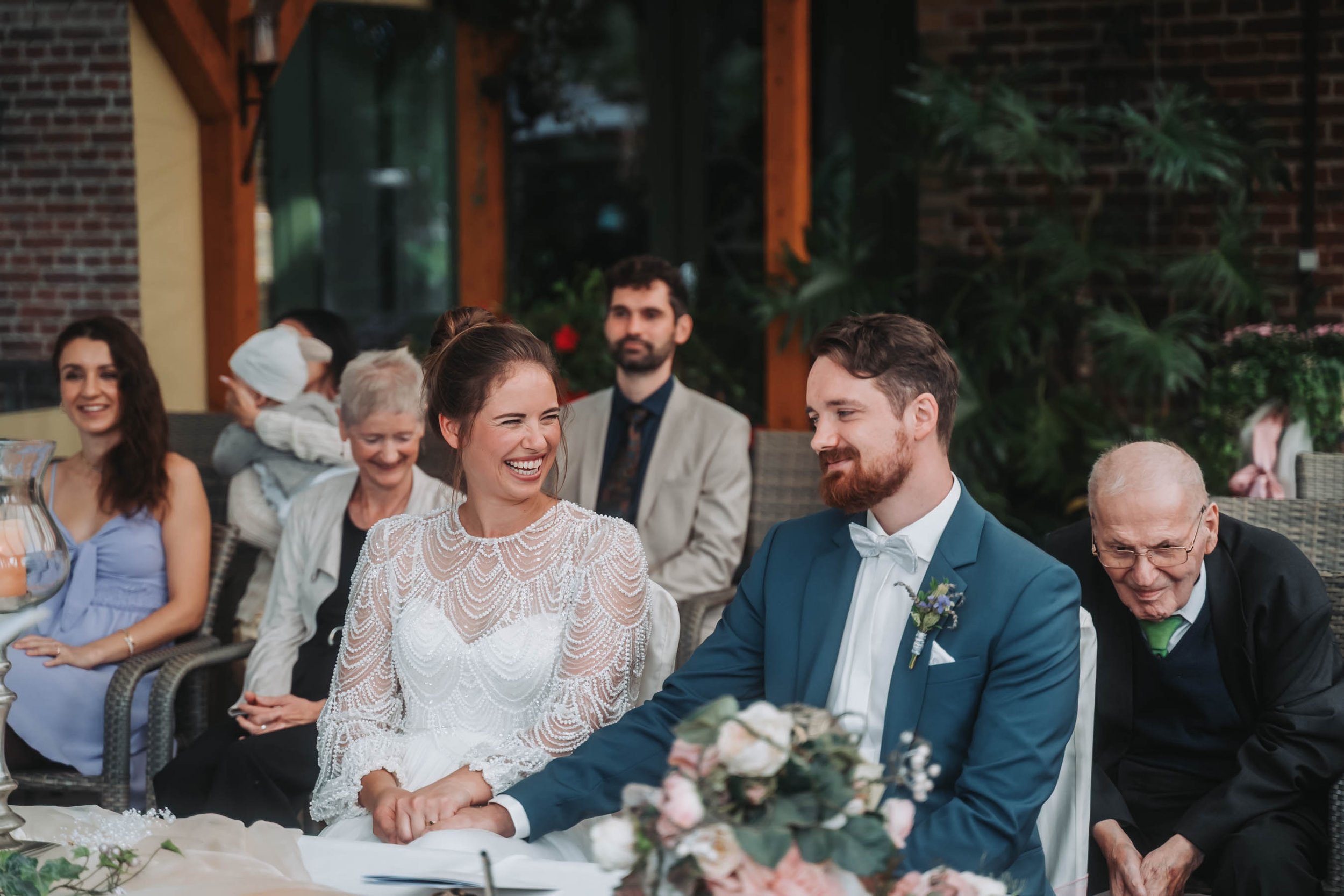 A bride and groom holding hands during their wedding ceremony, smiling at each other, surrounded by family and friends in an outdoor setting.