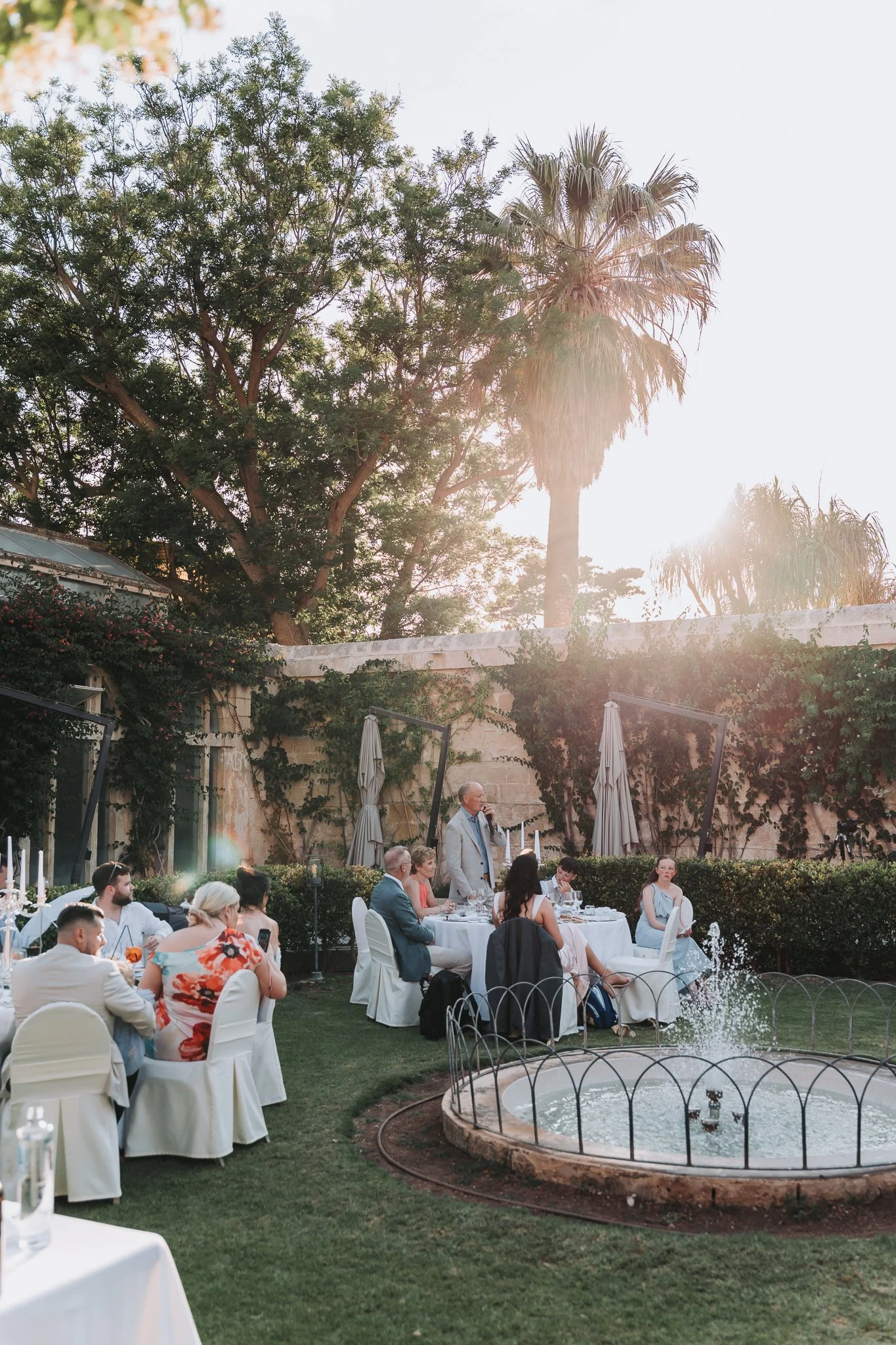 An outdoor wedding reception with guests seated at round tables under a tree, a man is giving a speech, surrounded by greenery, a fountain, and large trees with the sun shining through.