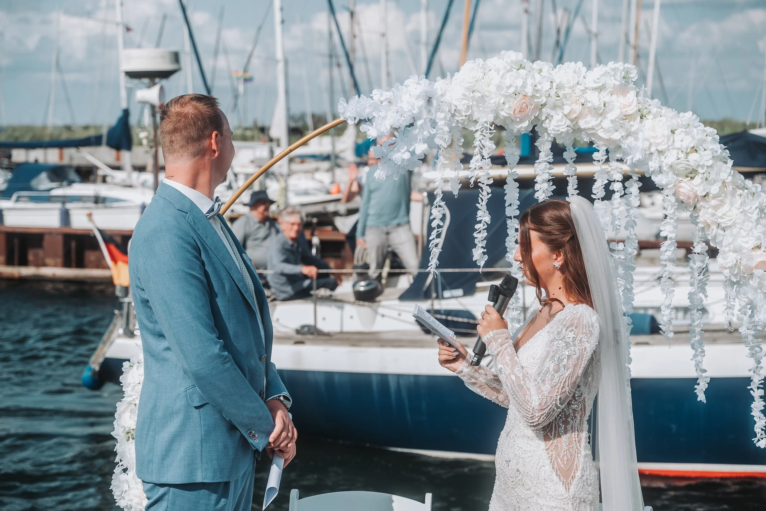 A bride and groom stand under a floral wedding arch near a marina with sailboats. The bride is holding a microphone and reading from a paper, while the groom is looking at her.
