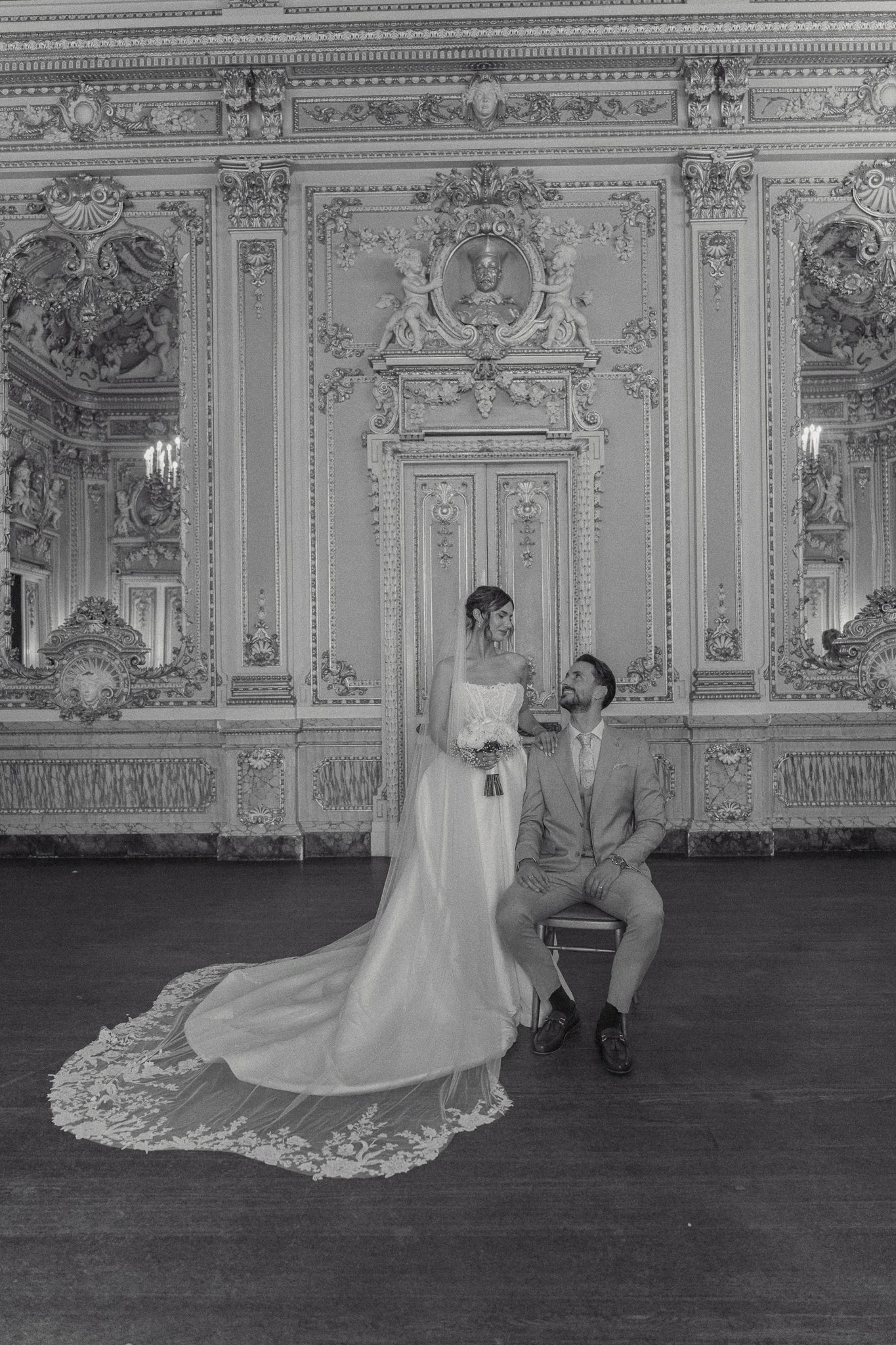 Black and white photo of a bride and groom in a grand, ornate room. The bride is standing in a wedding gown with a long train, holding a bouquet, while the groom is seated on a chair, dressed in a suit, looking up at her. The ornate room has detailed