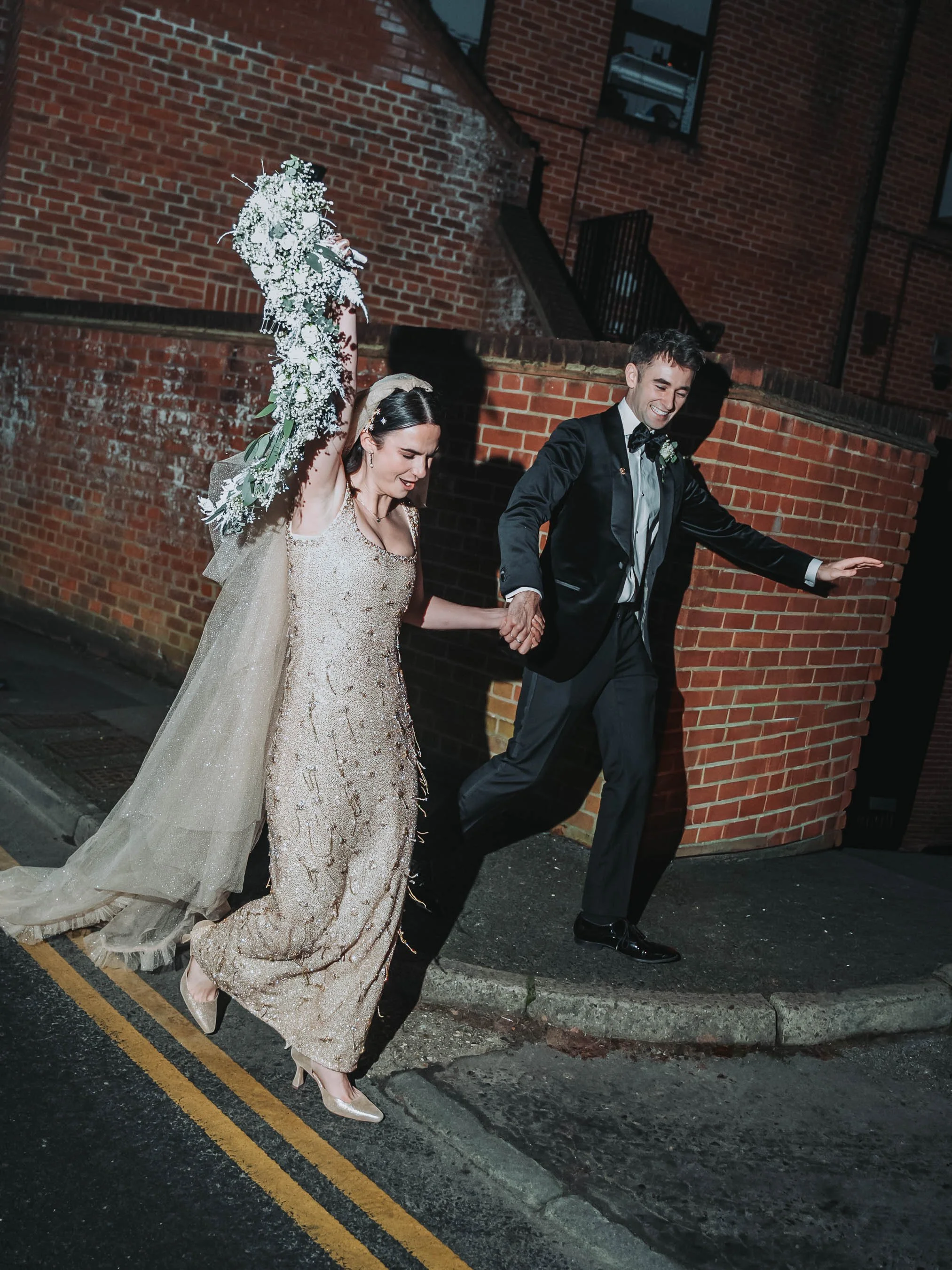 A bride and groom holding hands and walking on the street at night. The bride is wearing a sparkling gold dress with a veil and holding a bouquet of white flowers. The groom is dressed in a black tuxedo with a bow tie. They are smiling and appear joy