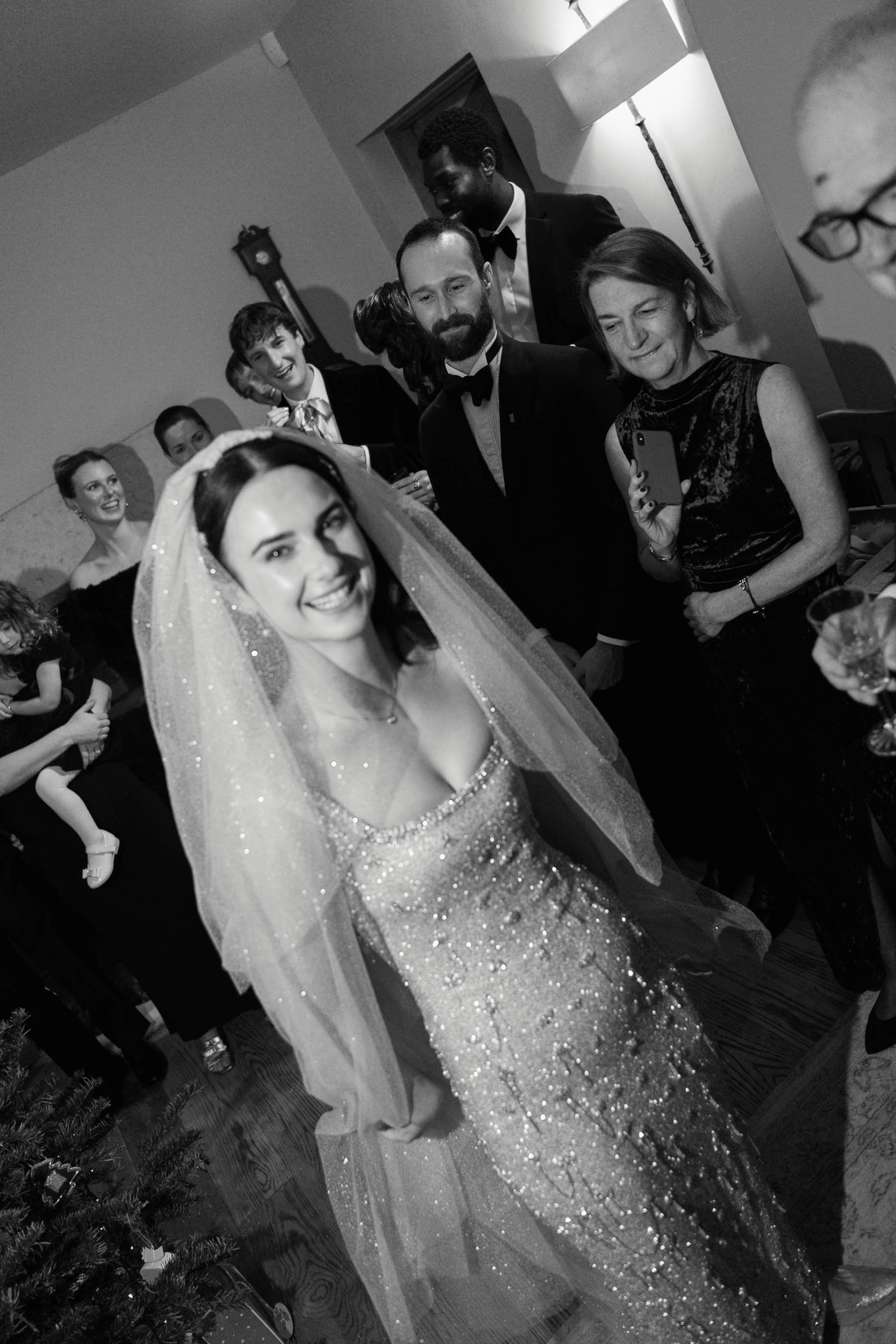 A black and white photo of a group of people at a party or celebration. A woman in a sparkly dress and veil is in the foreground, smiling. Behind her, several people in formal attire, including suits and dresses, are gathered, some smiling, some look