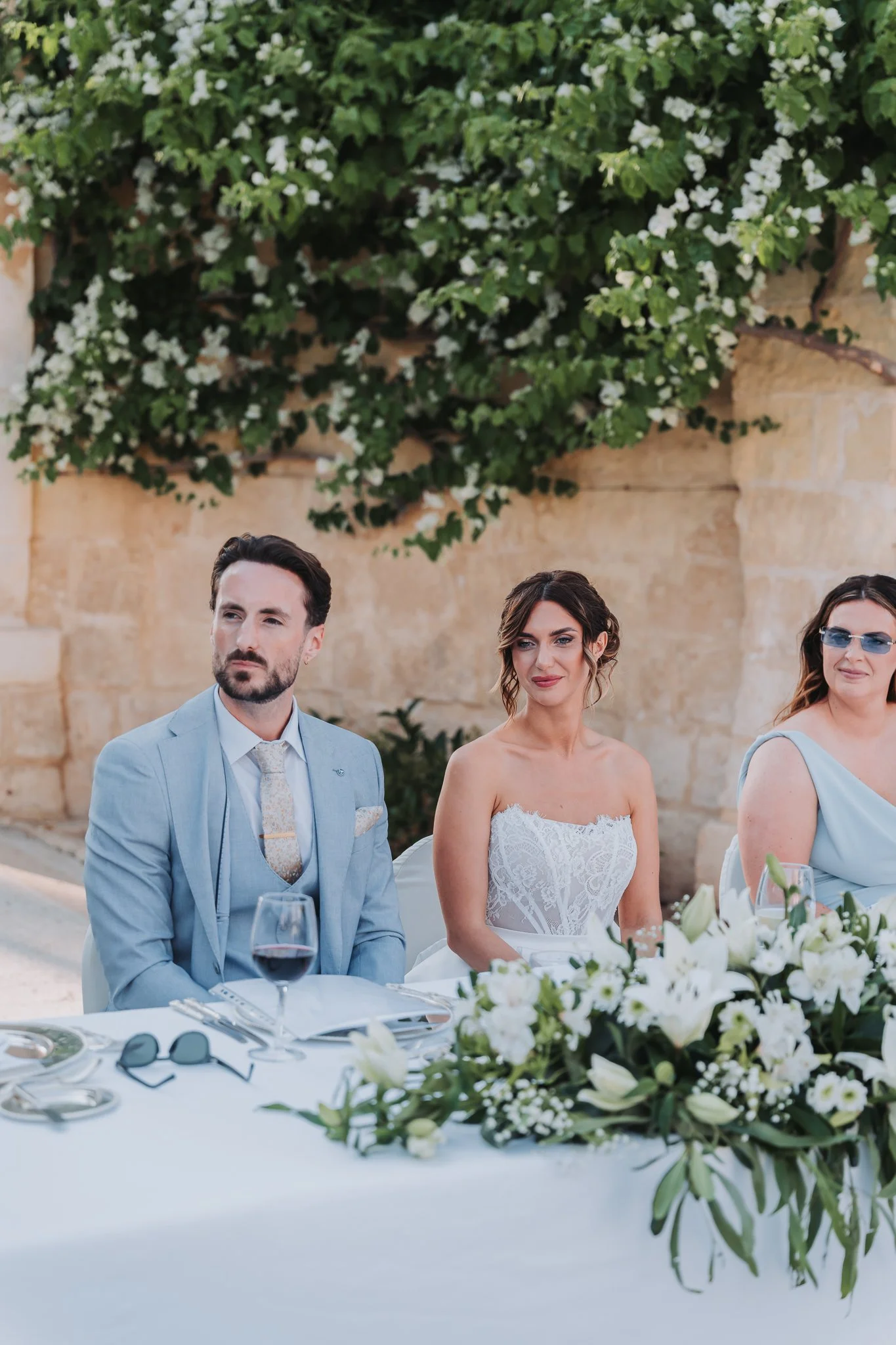 People attending a wedding reception sitting at a decorated table with floral arrangements, wine glasses, and tableware. A man in a light blue suit and a woman in a white strapless lace dress are visible.
