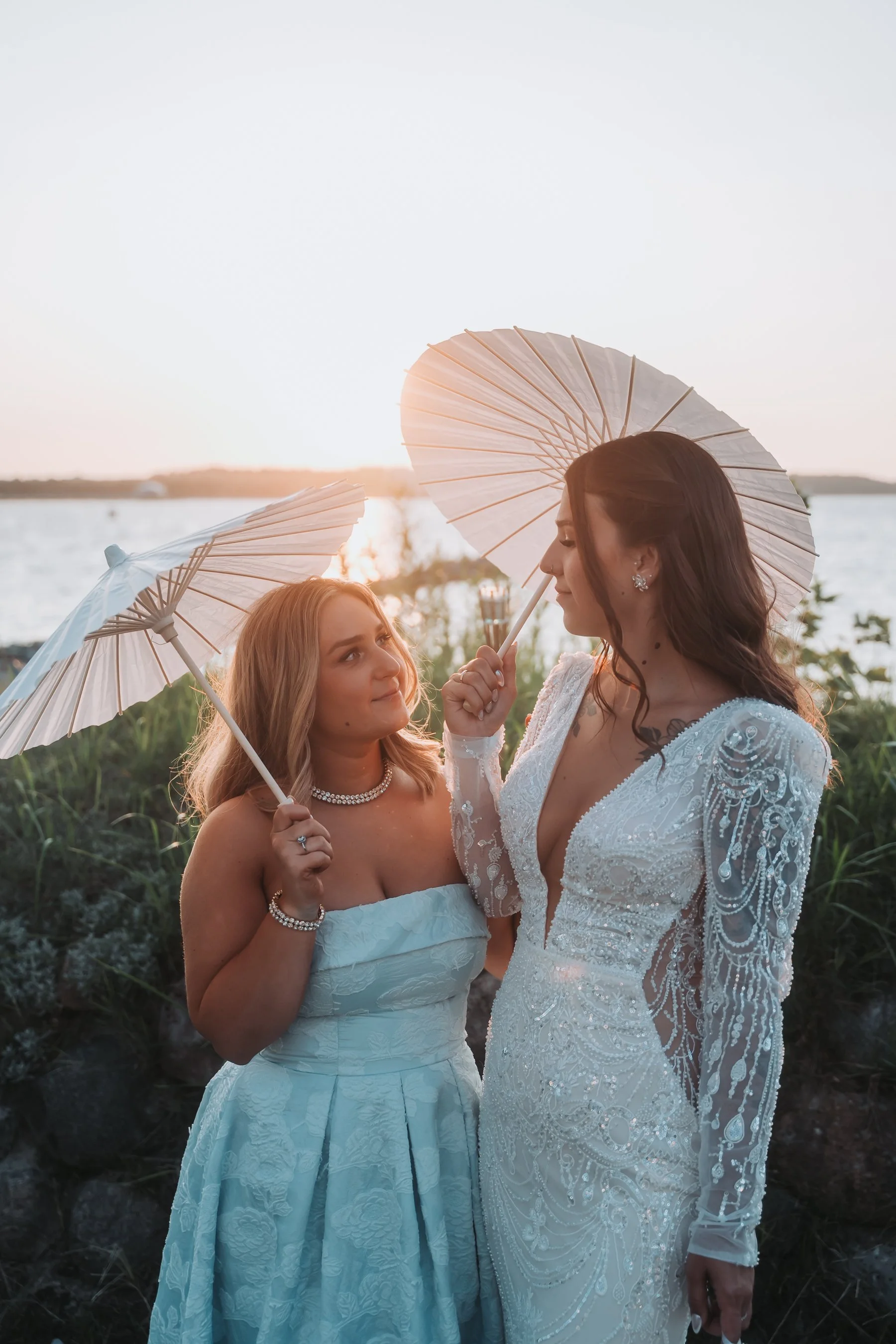 Two women dressed in wedding gowns holding lace parasols outdoors during sunset