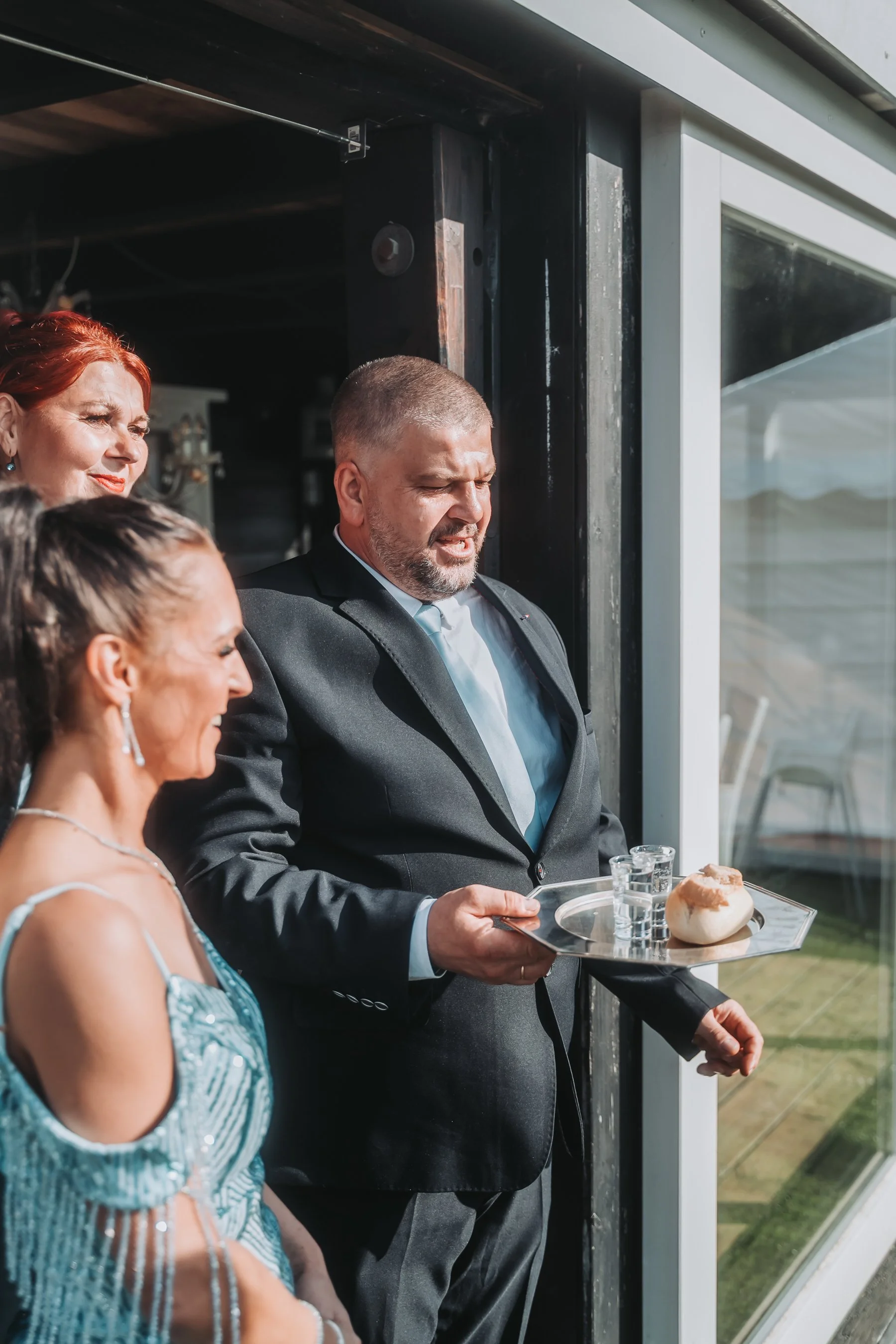 A man in a tuxedo holding a tray with drinks and bread rolls, standing next to a woman in a blue dress, with other people partially visible in the foreground, near a glass window.