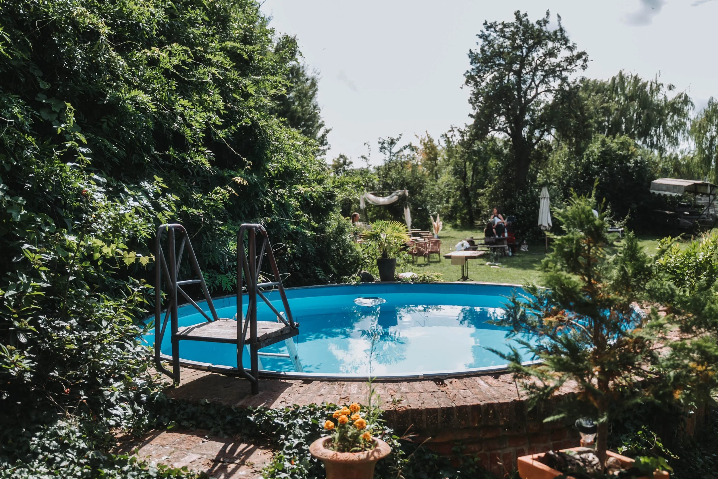 Backyard with a small above-ground pool, surrounded by greenery, outdoor chairs, and a table with umbrellas.