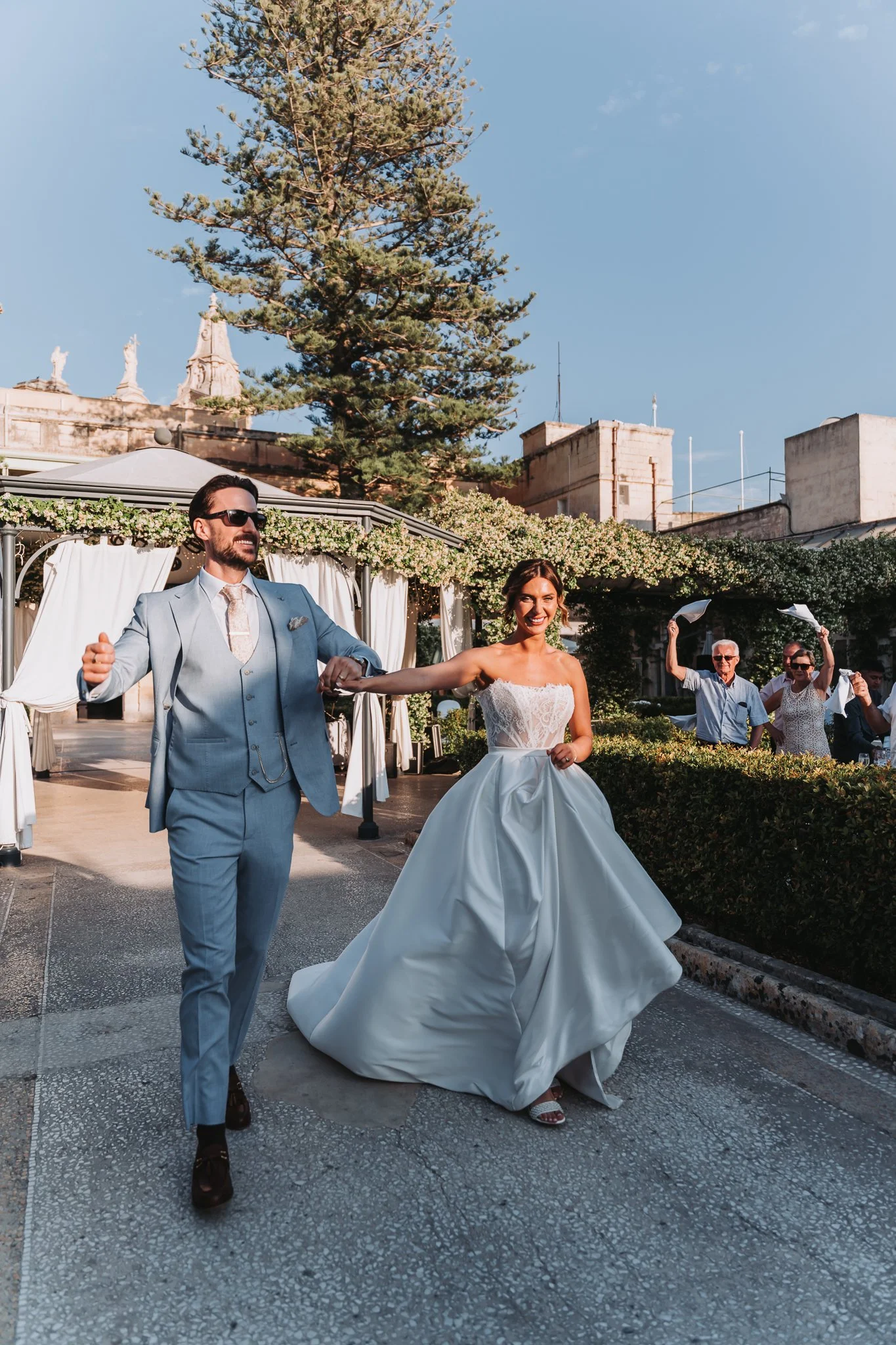 A newlywed couple is dancing outdoors during their wedding celebration. The bride is wearing a strapless white wedding gown and the groom is in a light blue suit with sunglasses. They are smiling and holding hands, with older guests in the background