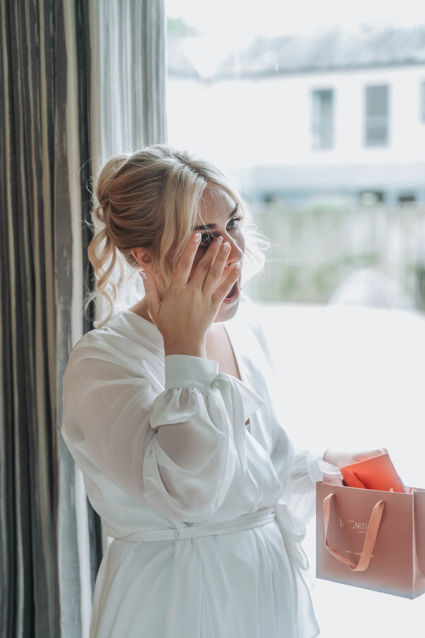 A woman with blonde hair in an updo, wearing a white dress with puffed sleeves, holds her hand to her face and appears surprised or excited, looking out a window with window panes behind her, holding a pink shopping bag.