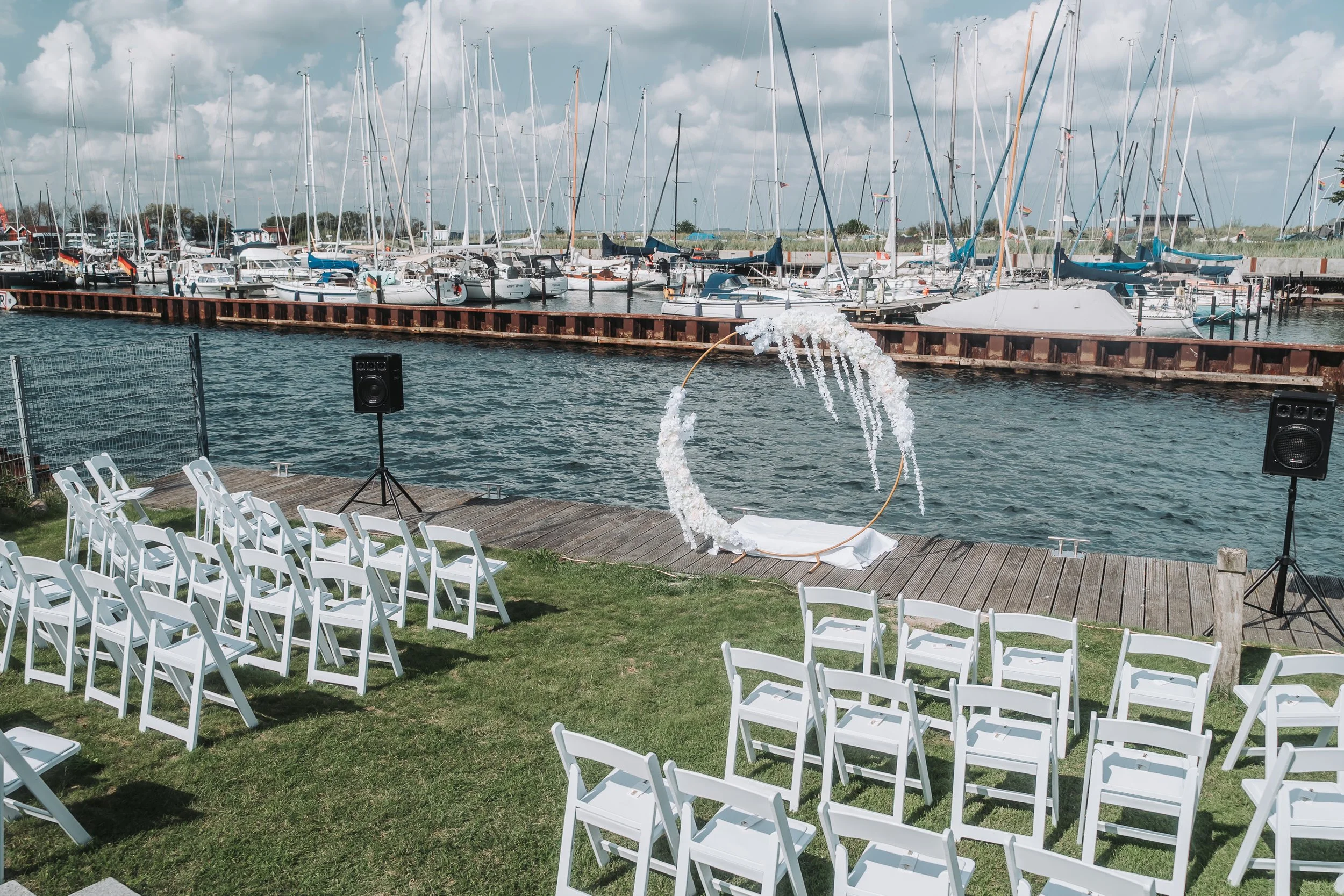 A marina with sailboats docked along a pier under a partly cloudy sky. In the foreground, there are white chairs set up on a grassy area facing the water, and a decorative hoop with white flowers and fabric, suggesting a wedding or event setup.