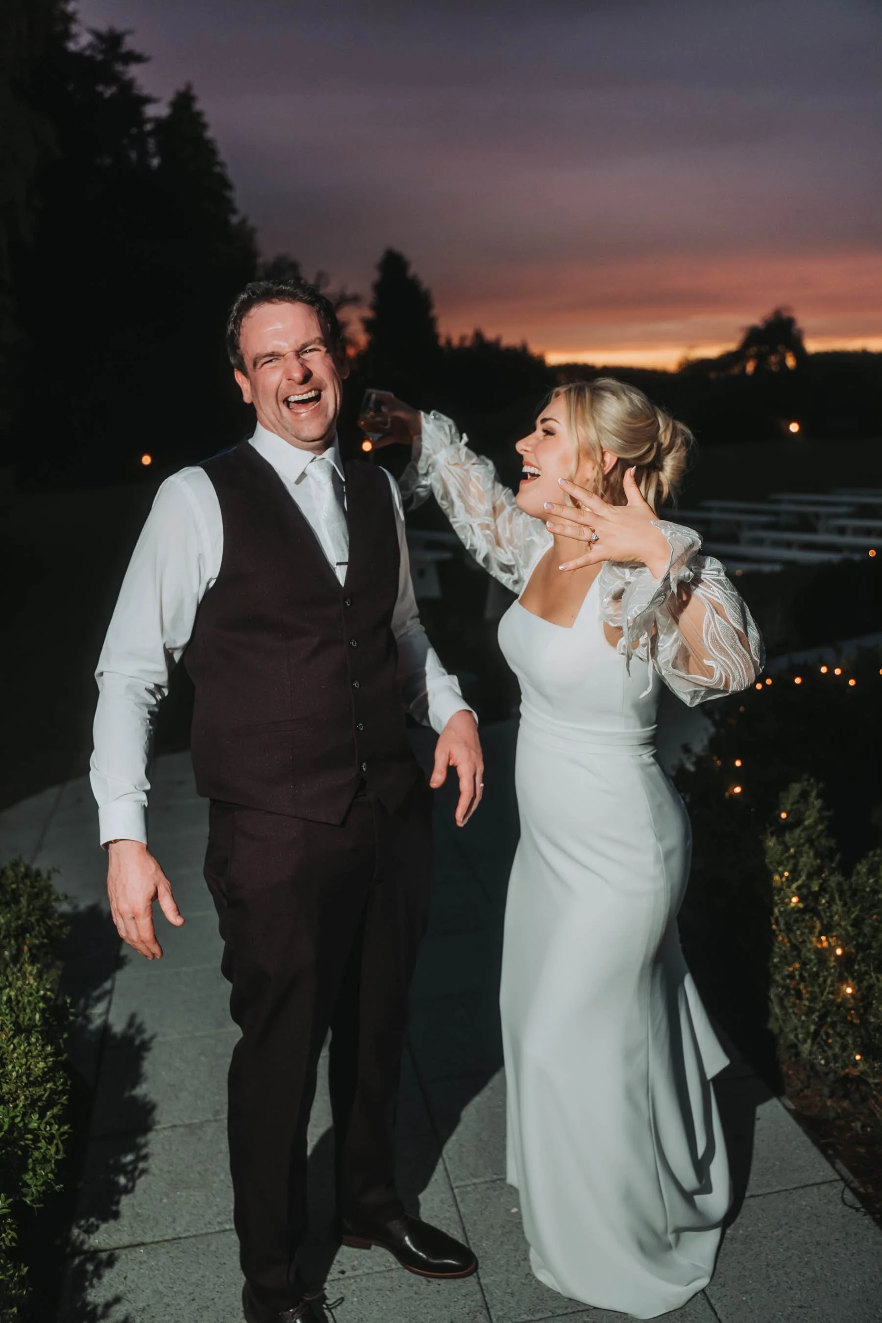 A man and a woman in formal wedding attire laughing and enjoying each other's company outdoors during sunset, with trees and decorative lights in the background.