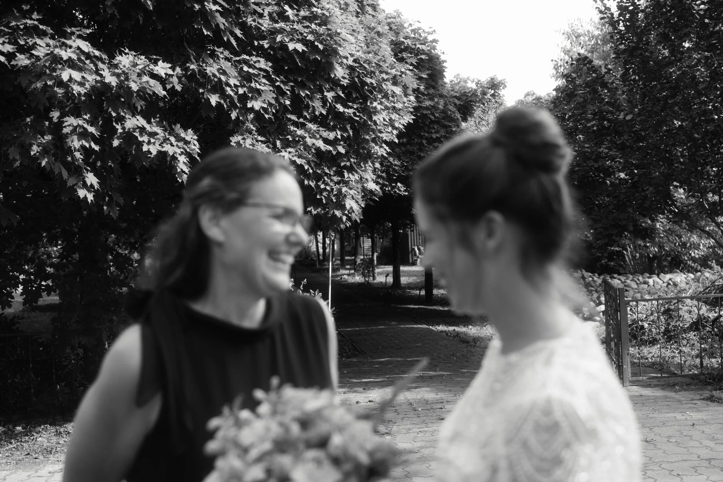 Black and white photo of two young women smiling and talking outdoors, with trees and a paved path in the background. One is wearing glasses, and the other has her hair in a bun.