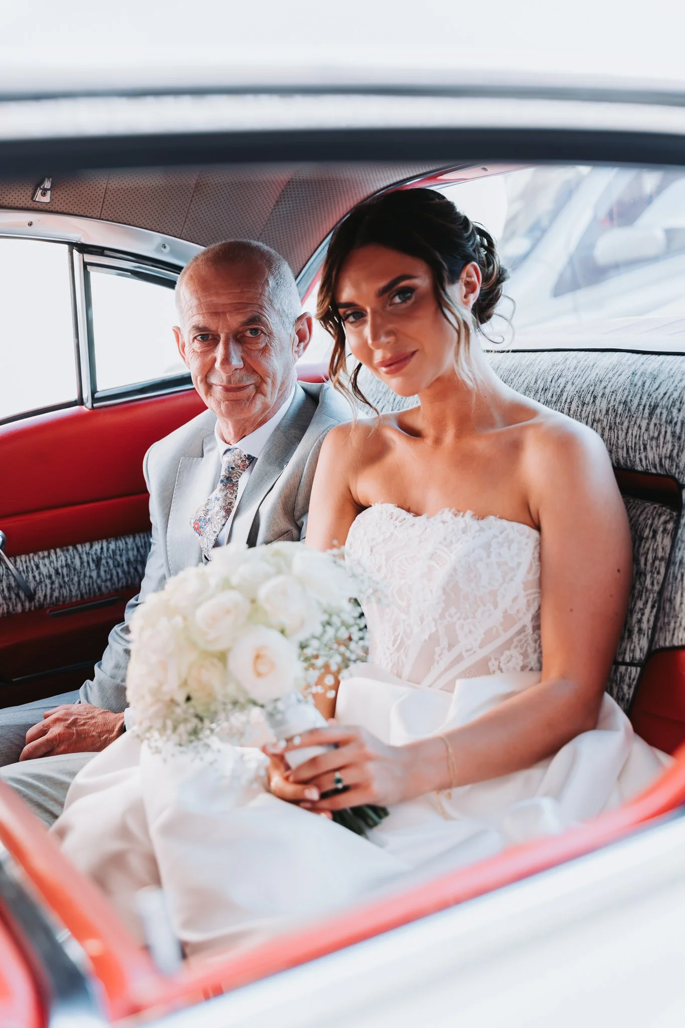 A bride and an elderly man, likely her father, sitting inside a vintage car on her wedding day. The bride is holding a bouquet of white flowers and wearing a white lace wedding dress. Both are looking at the camera.