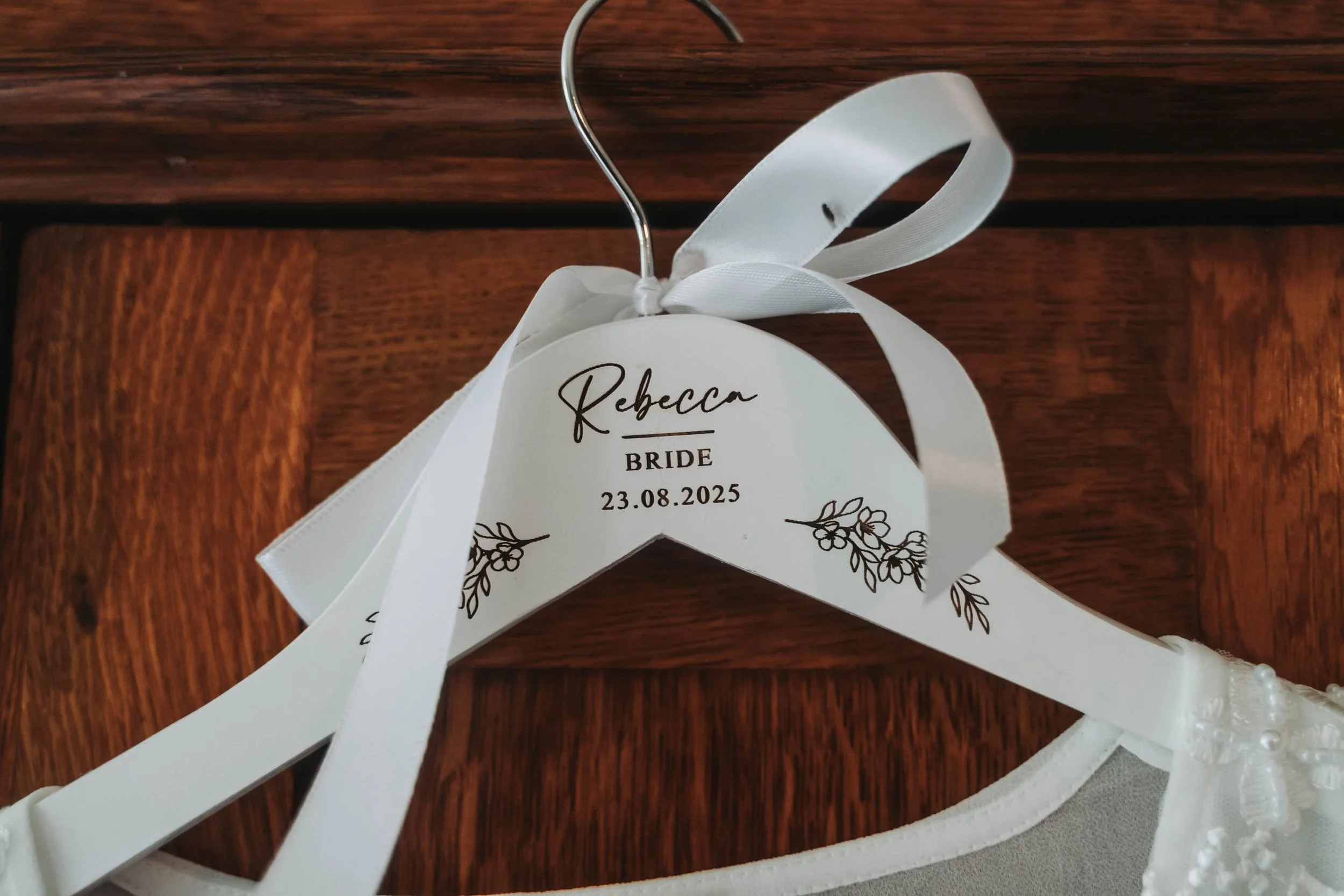 A white wedding or special occasion decorative sign with the name 'Rebecca,' the date 23.08.2025, and floral illustrations, attached to a white lace and pearl headband with a ribbon, arranged on a dark wooden surface.