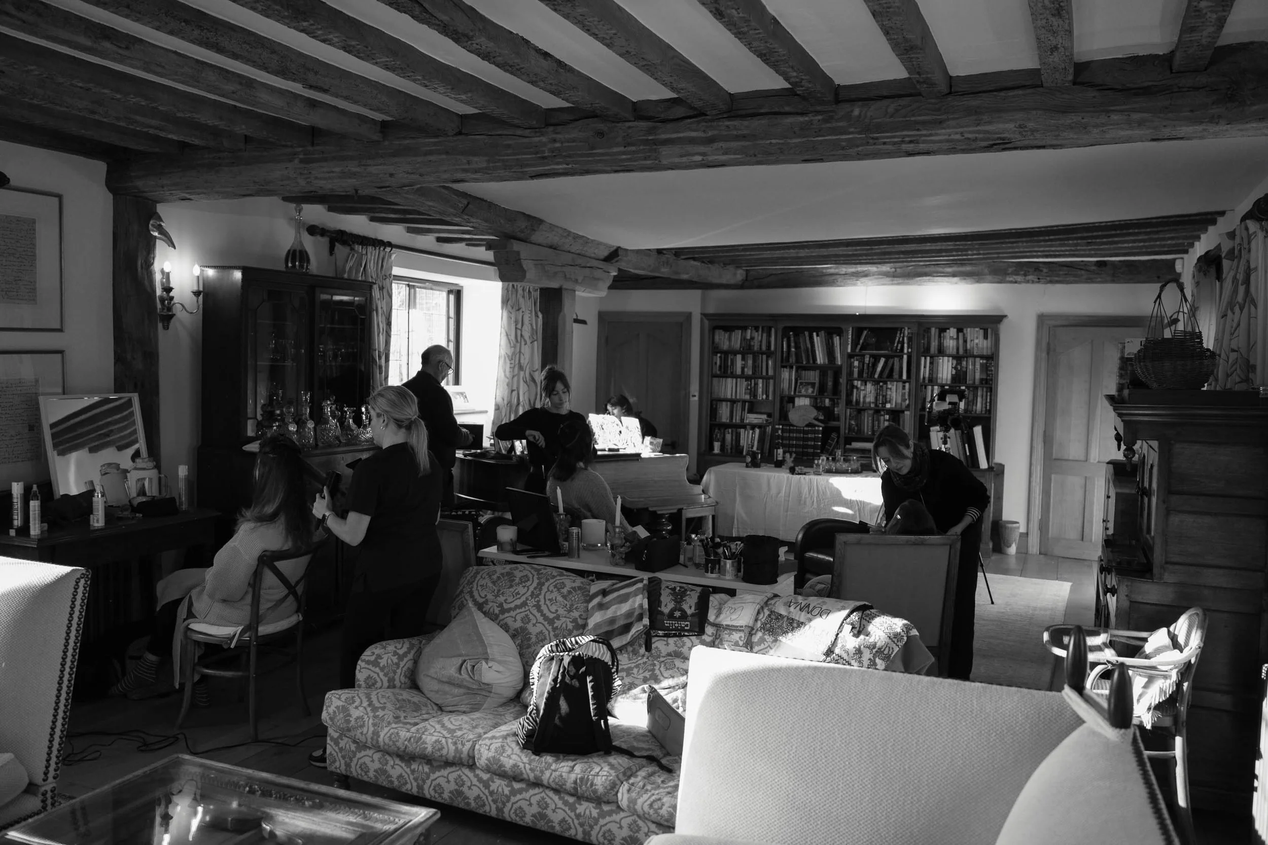 A cozy living room with wooden beams on the ceiling and a bookshelf filled with books on the wall. Several people are gathered, some sitting and some standing, engaged in various activities such as reading, talking, or working on laptops. There is a 
