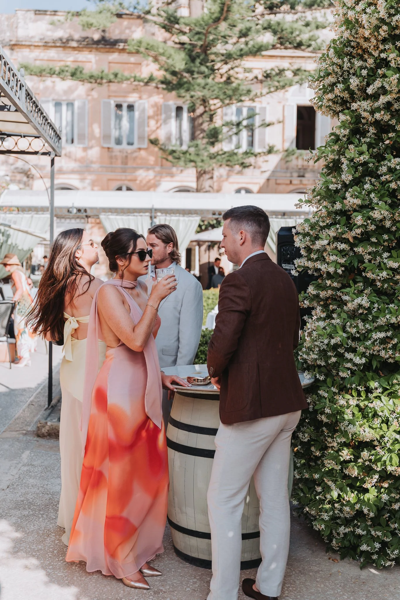 Group of people socializing at an outdoor event, standing around a barrel table with drinks, amidst greenery and a building in the background.