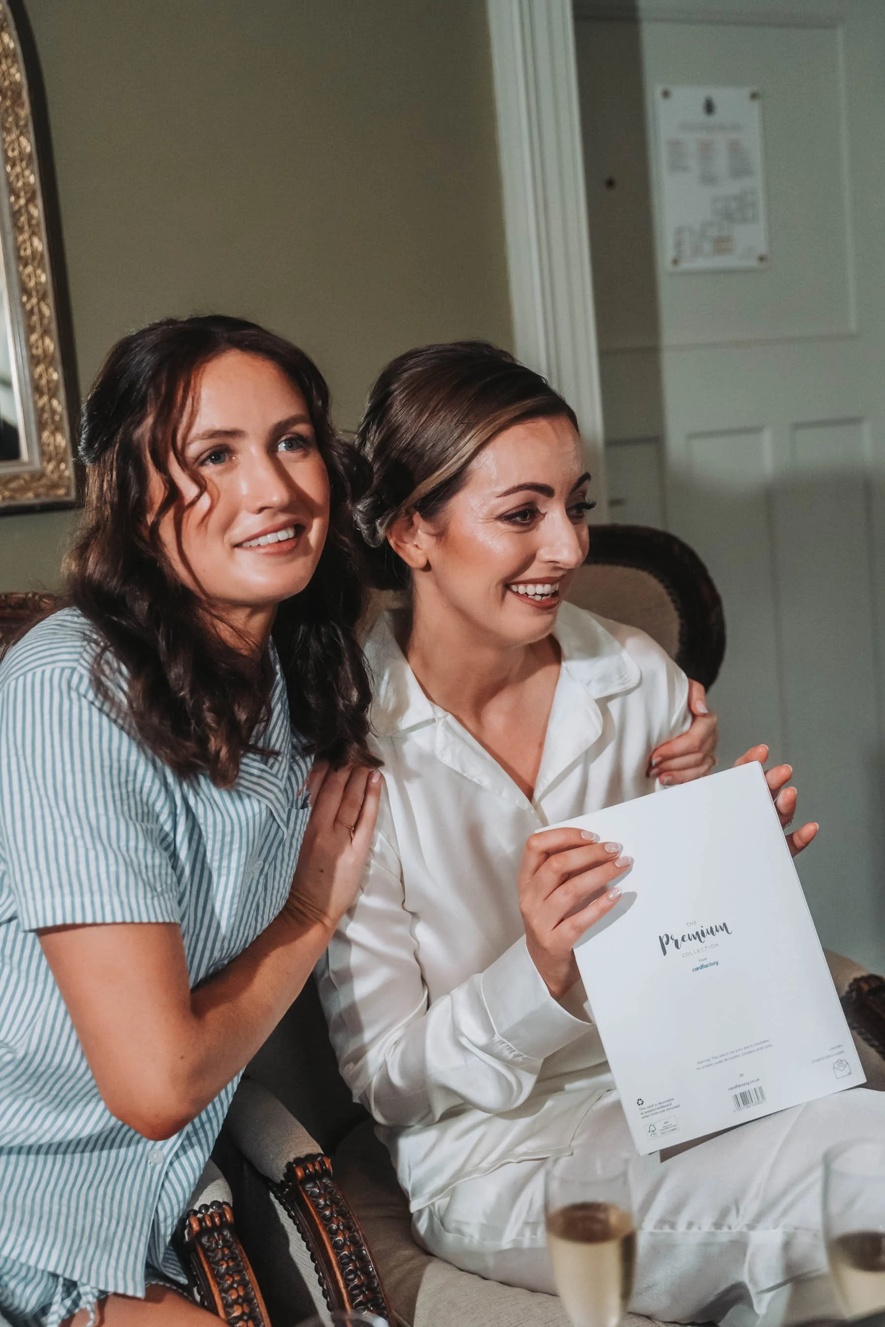 Two women sitting together, smiling. One woman is holding a white booklet or menu that reads 'The Premium Collection.' They are sitting indoors with a green wall and a door visible in the background.