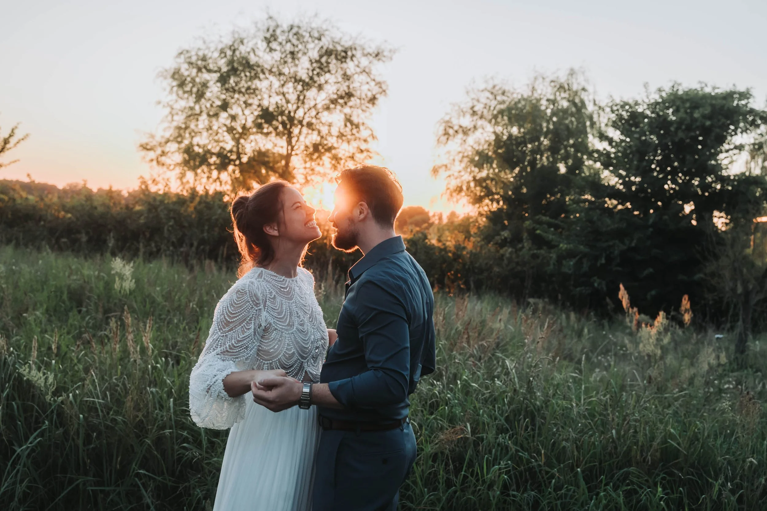 A couple stands outdoors in a grassy field at sunset, smiling and holding hands while gazing at each other.