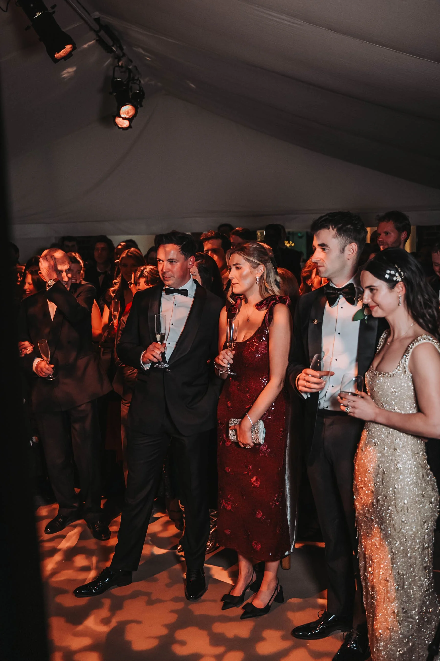 A group of men and women dressed in formal evening wear, standing under a tent with decorative lighting, holding wine glasses and looking engaged in conversation at a social event or celebration.