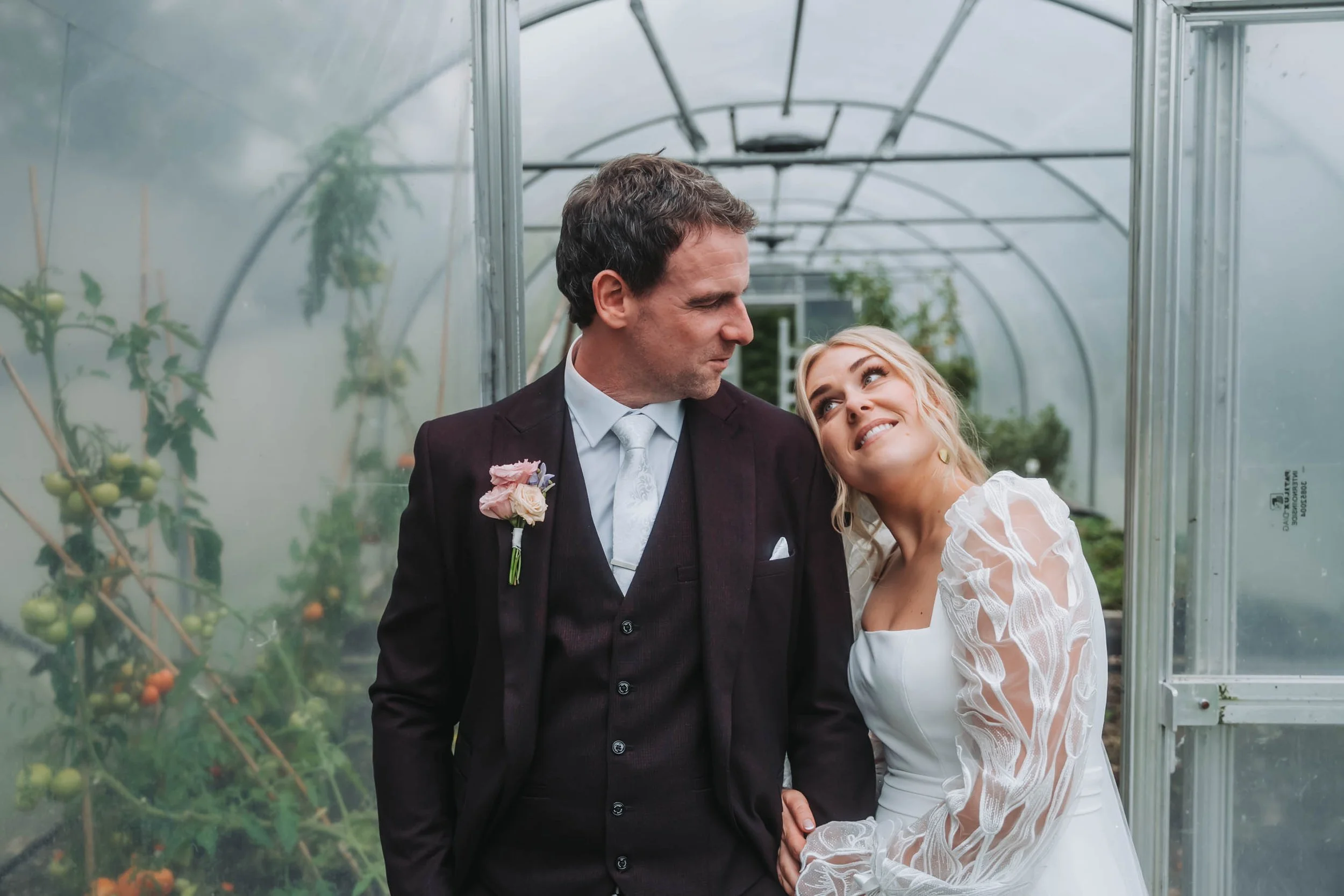A man and woman in wedding attire smiling and looking at each other inside a greenhouse with tomato plants in the background.