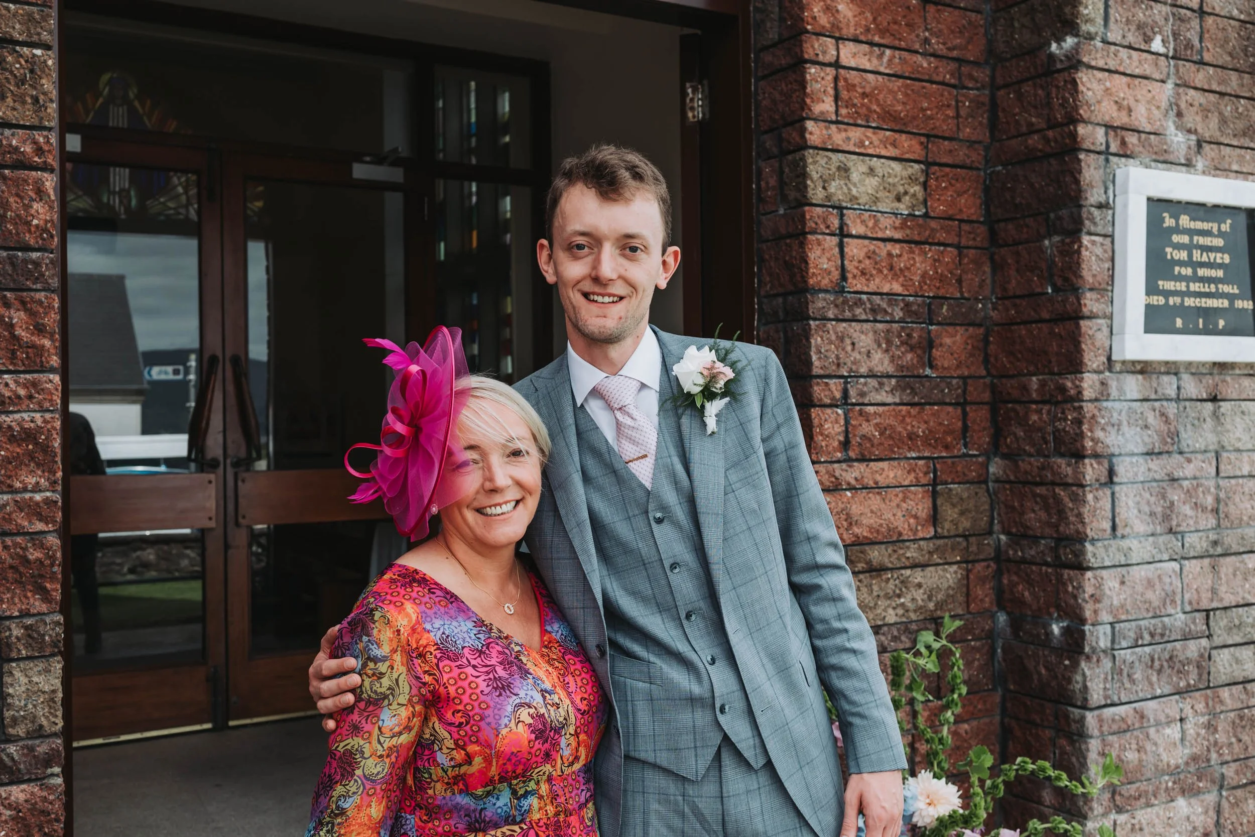 A young man in a gray suit with a boutonniere stands next to an older woman wearing a colorful dress and large pink fascinator. They are smiling and standing outside a brick building with a memorial plaque. They are embracing each other.