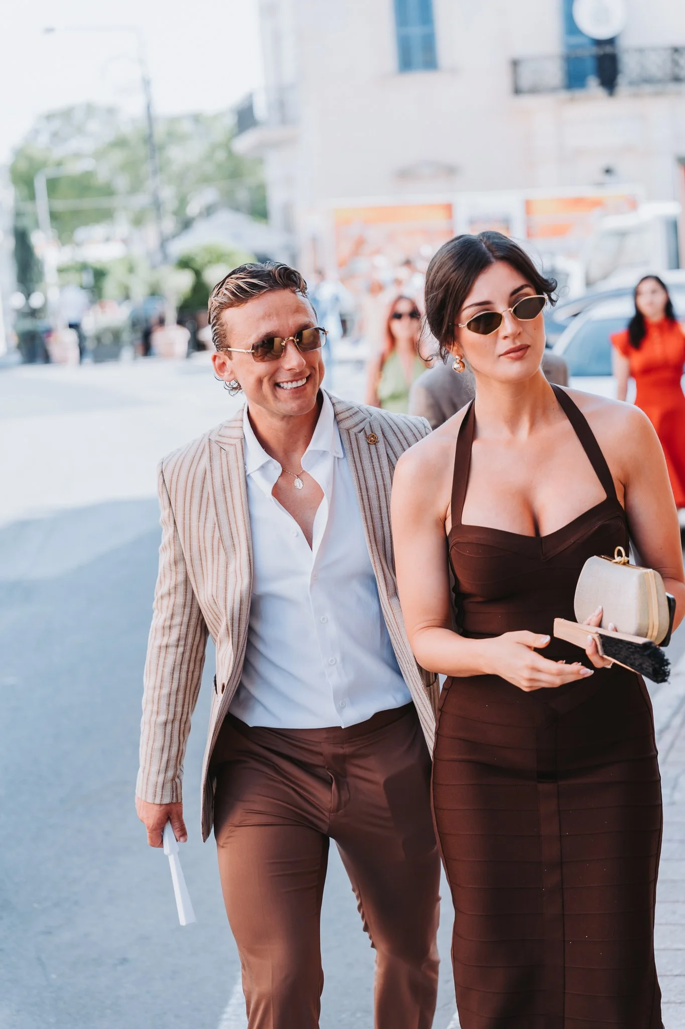 Two people wearing sunglasses walking on a city street, with others and buildings in the background.