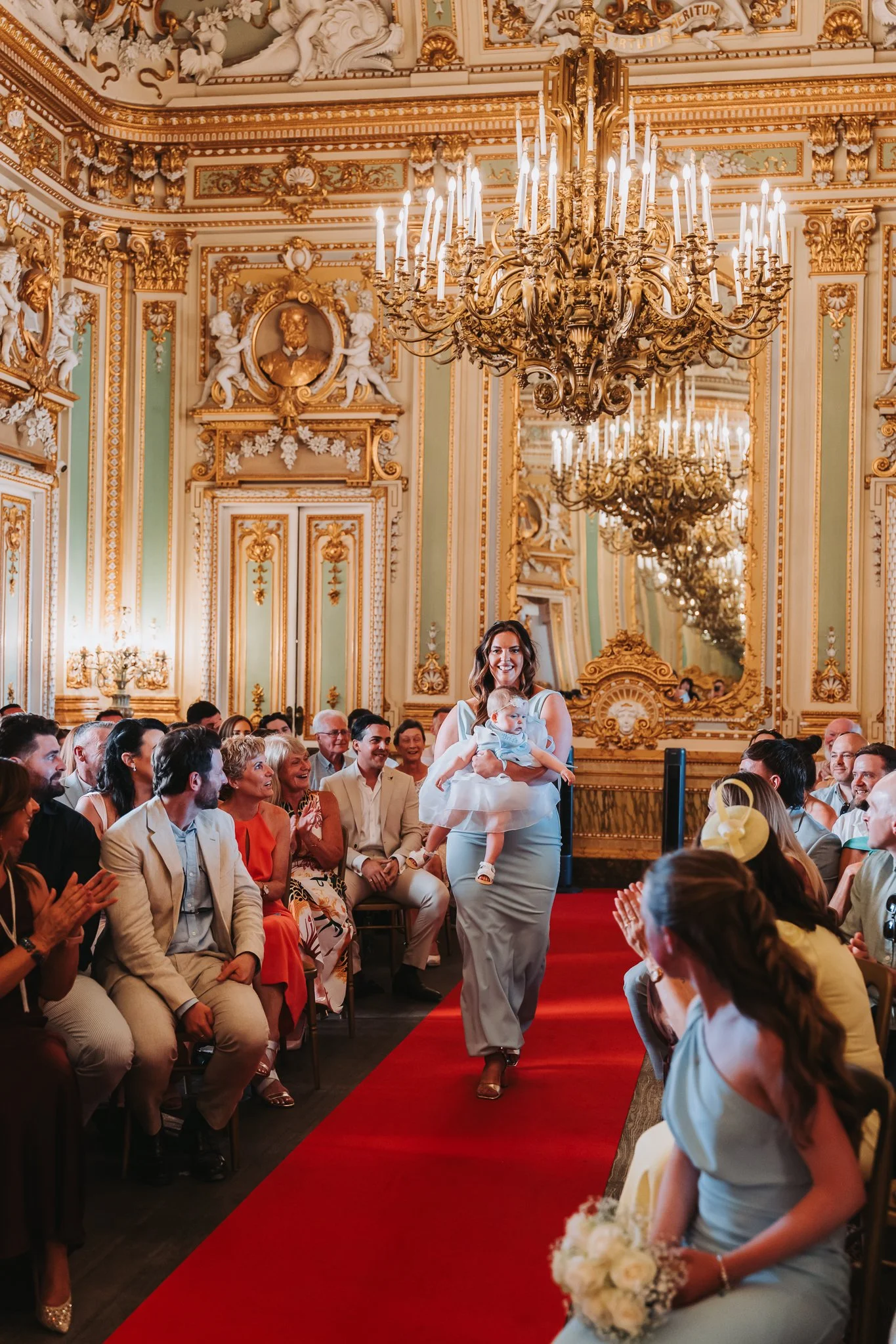 A woman in a white dress holding a baby walks down a red carpet in a lavishly decorated room with gold accents, chandeliers, and a large mirror. Guests are seated on either side, clapping and smiling.